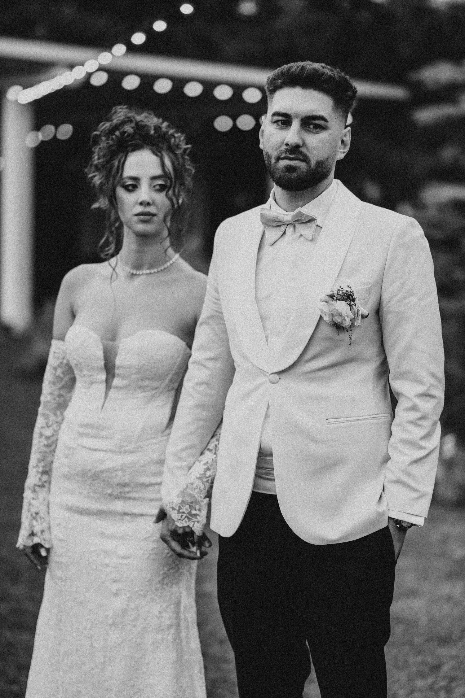 Black and white wedding photo of bride in lace gown and groom in white tuxedo holding hands outdoors.