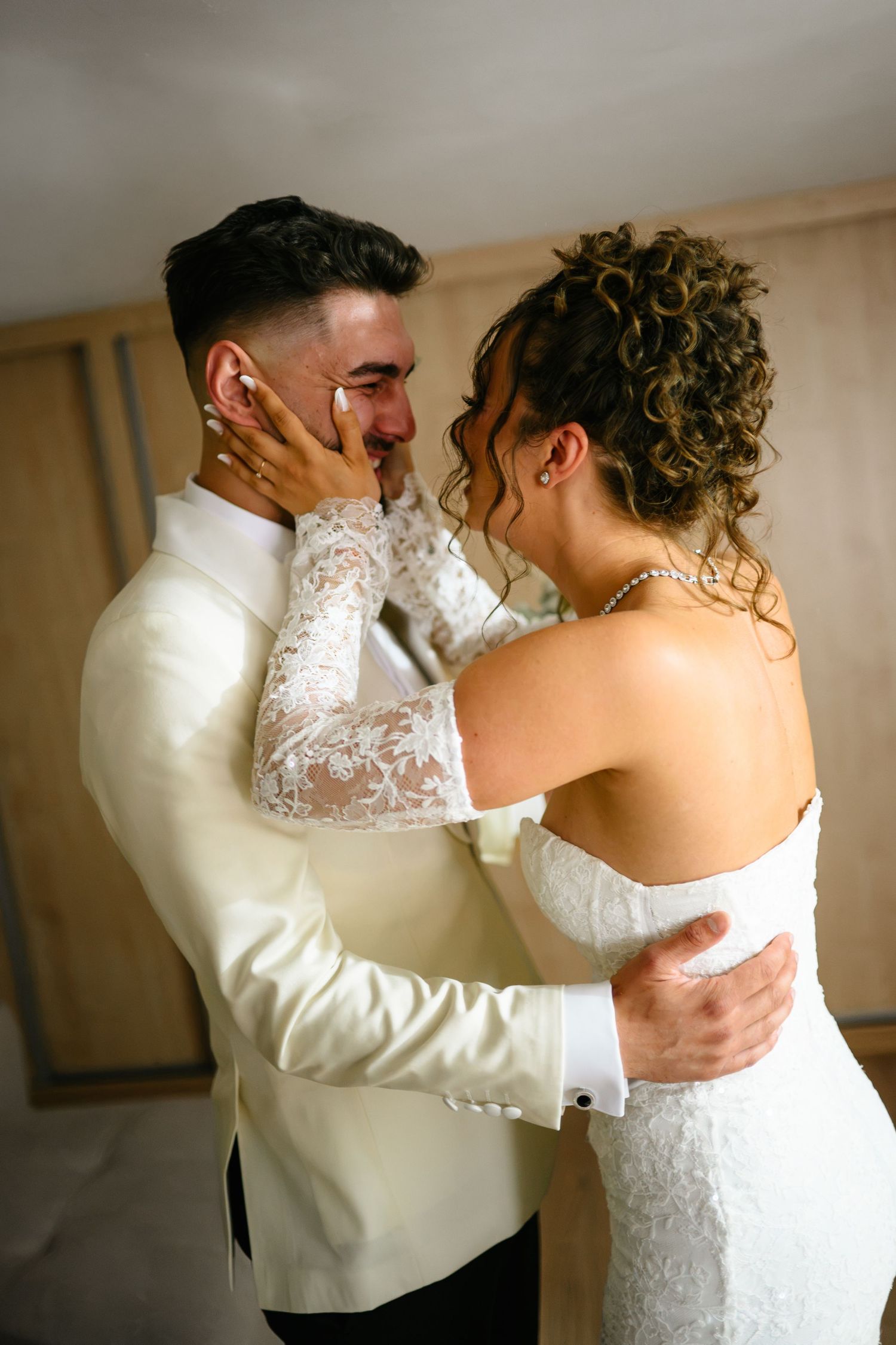 Emotional groom crying as bride tenderly cups his face during intimate wedding moment, both dressed in elegant white attire.