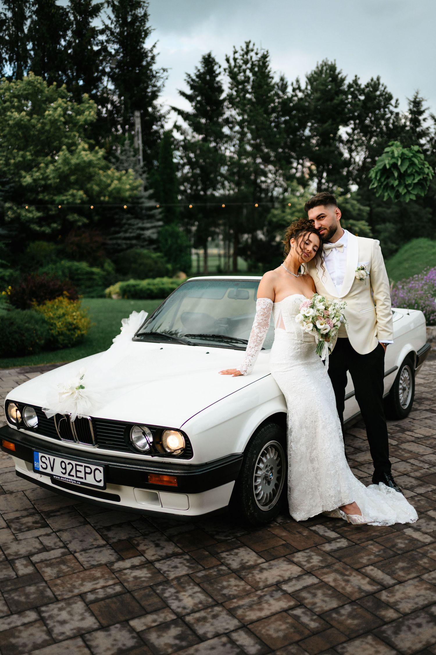 Bride and groom posing by a classic white BMW convertible decorated with ribbons on a cobblestone driveway.