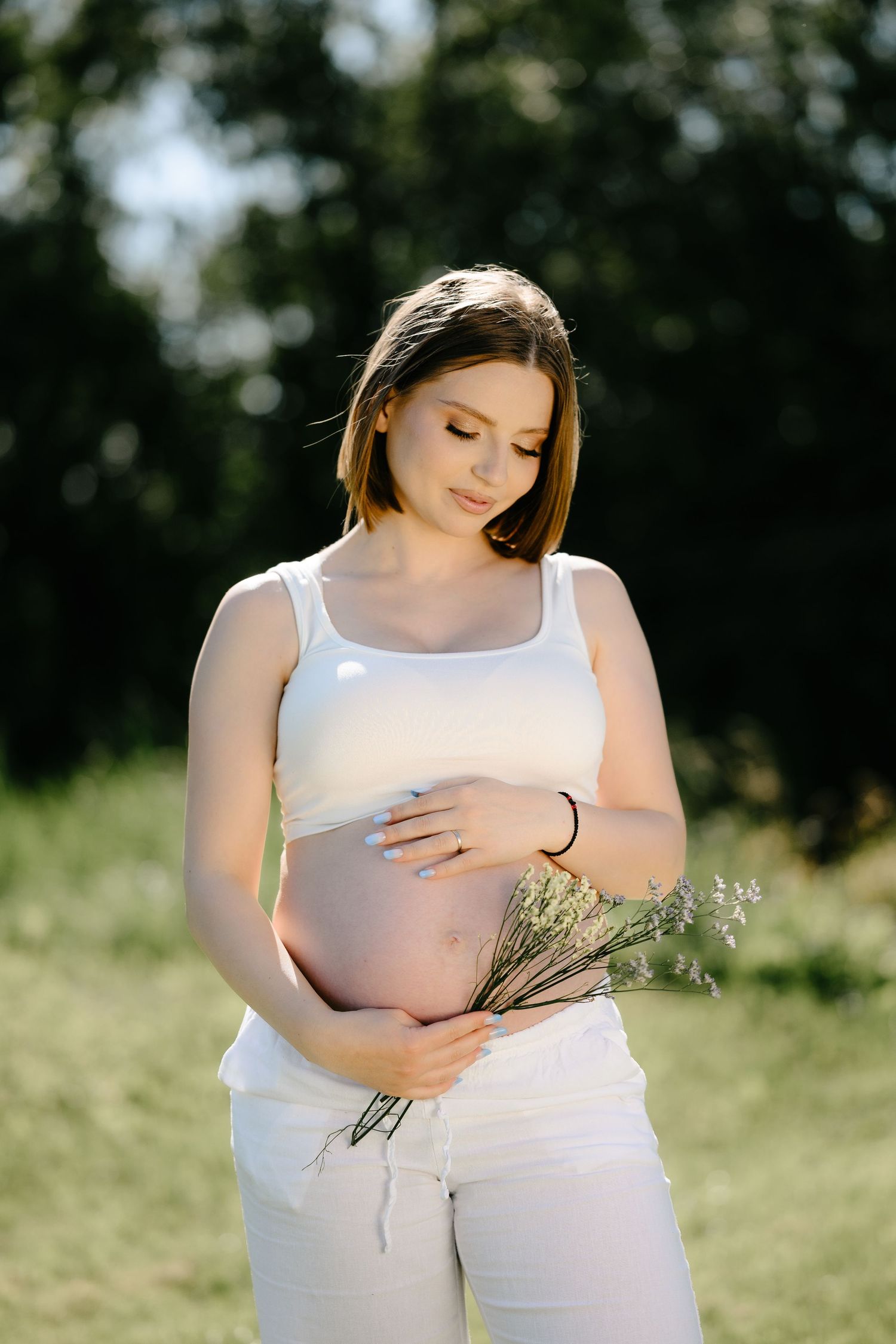 Pregnant woman in white outfit cradling baby bump while holding lavender bouquet in sunny garden.