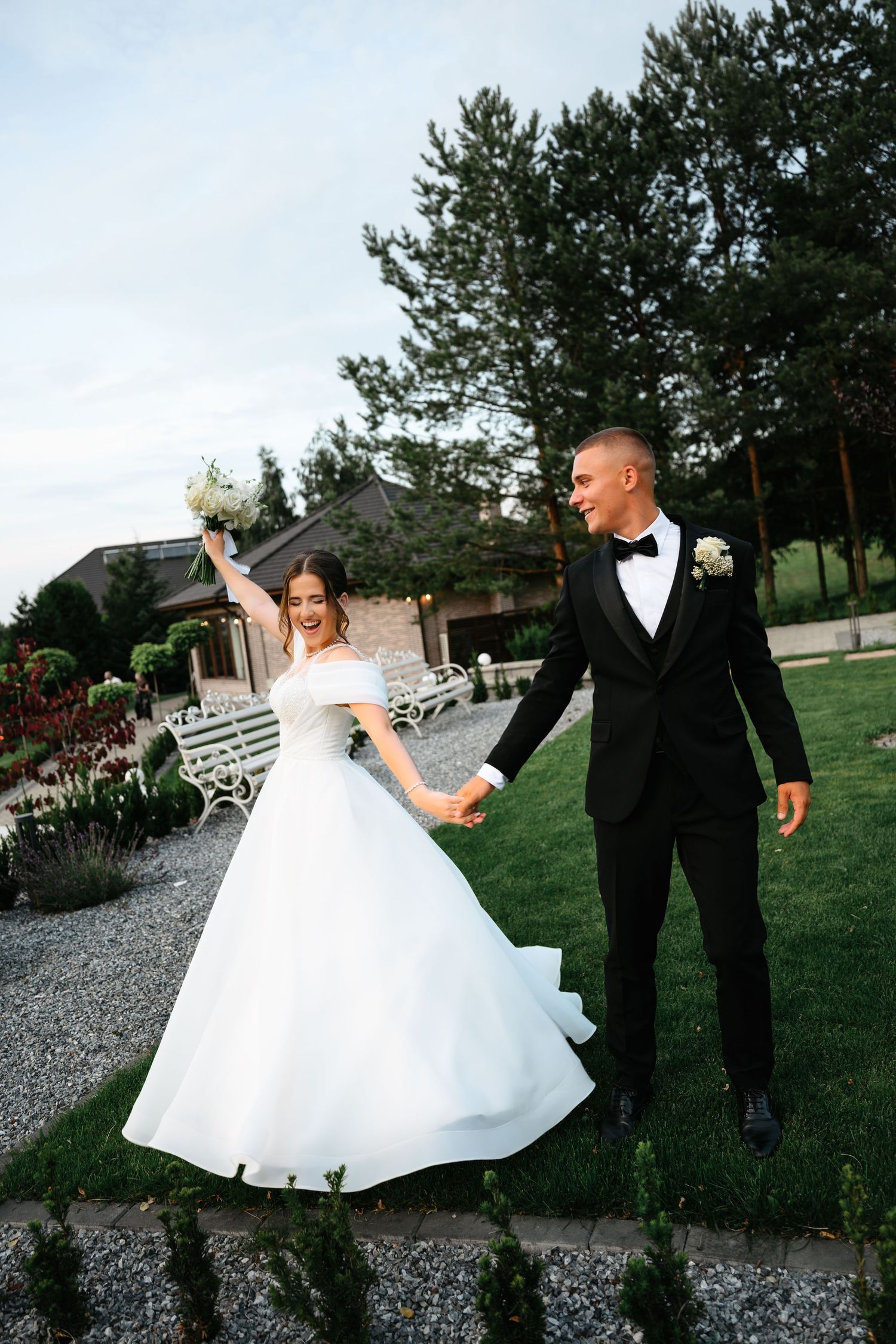 A joyful bride in a white gown and groom in a black tuxedo hold hands outdoors at a wedding venue.