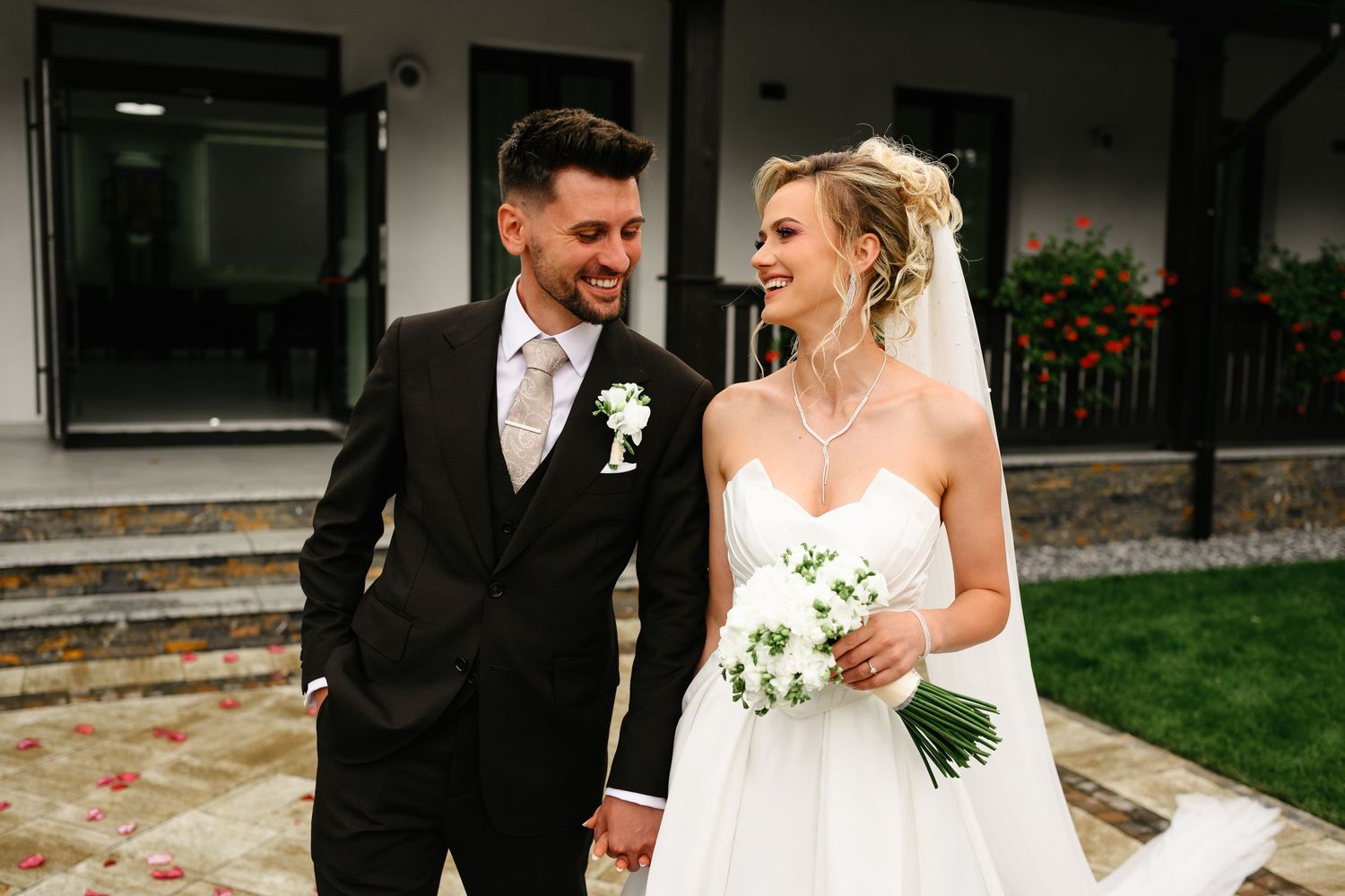 Happy newlywed couple walking hand in hand, bride in white gown with bouquet, groom in dark suit, outdoor venue.