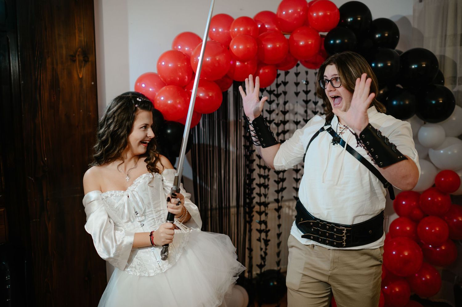 Woman in white bridal costume playfully holds sword near laughing guest at Halloween balloon party.