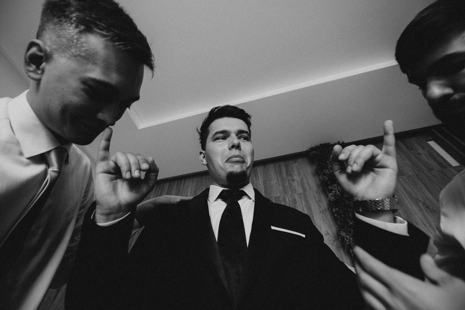 Black and white photo of groom in suit surrounded by groomsmen pointing fingers during wedding prep.