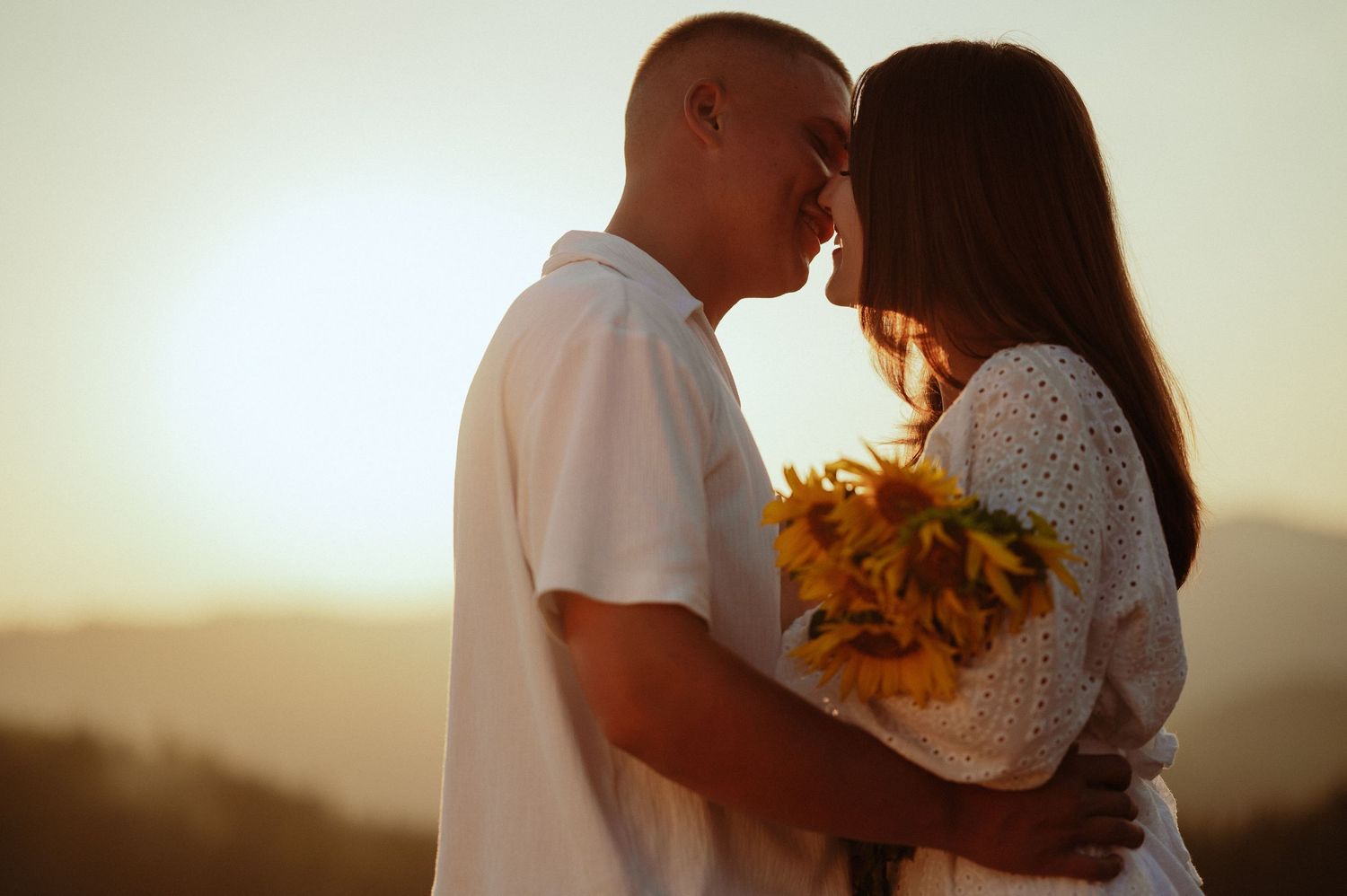 Couple embracing and almost kissing holding sunflowers in warm glowing sunset light.