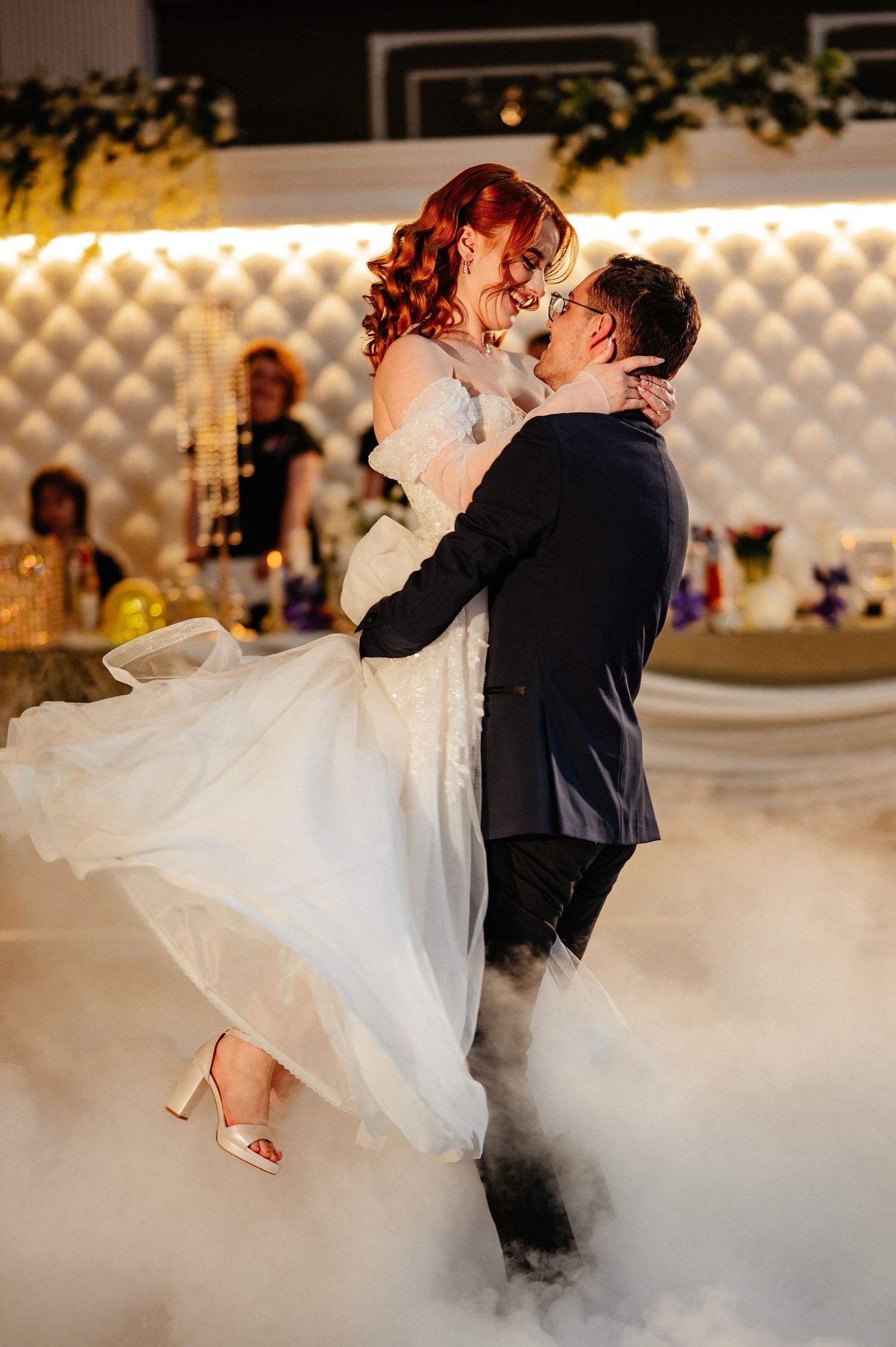 Groom lifts bride during romantic first dance surrounded by dramatic fog at wedding reception.