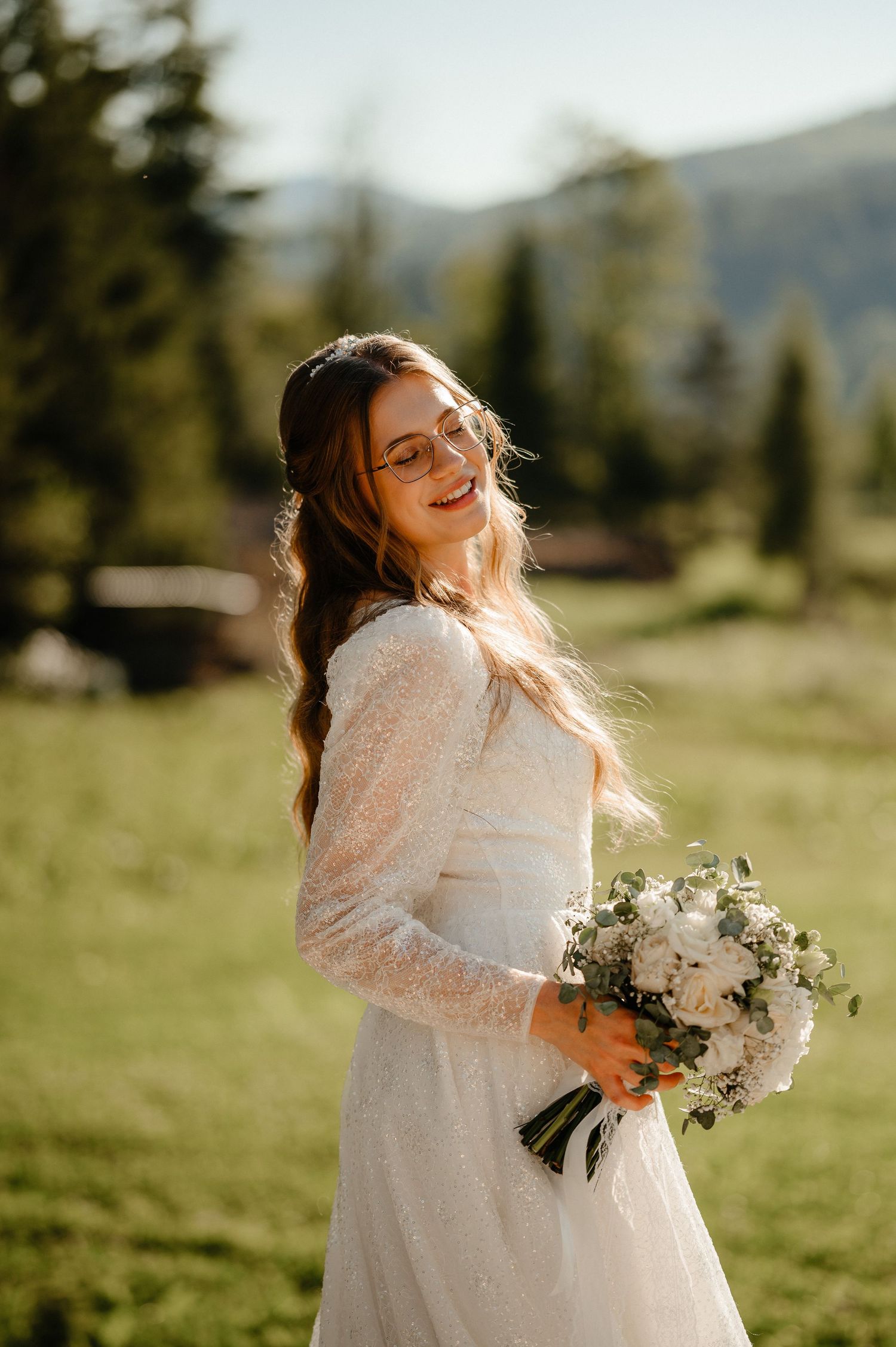 Smiling bride in sparkly long-sleeve wedding dress holding white bouquet in sunny mountain meadow.