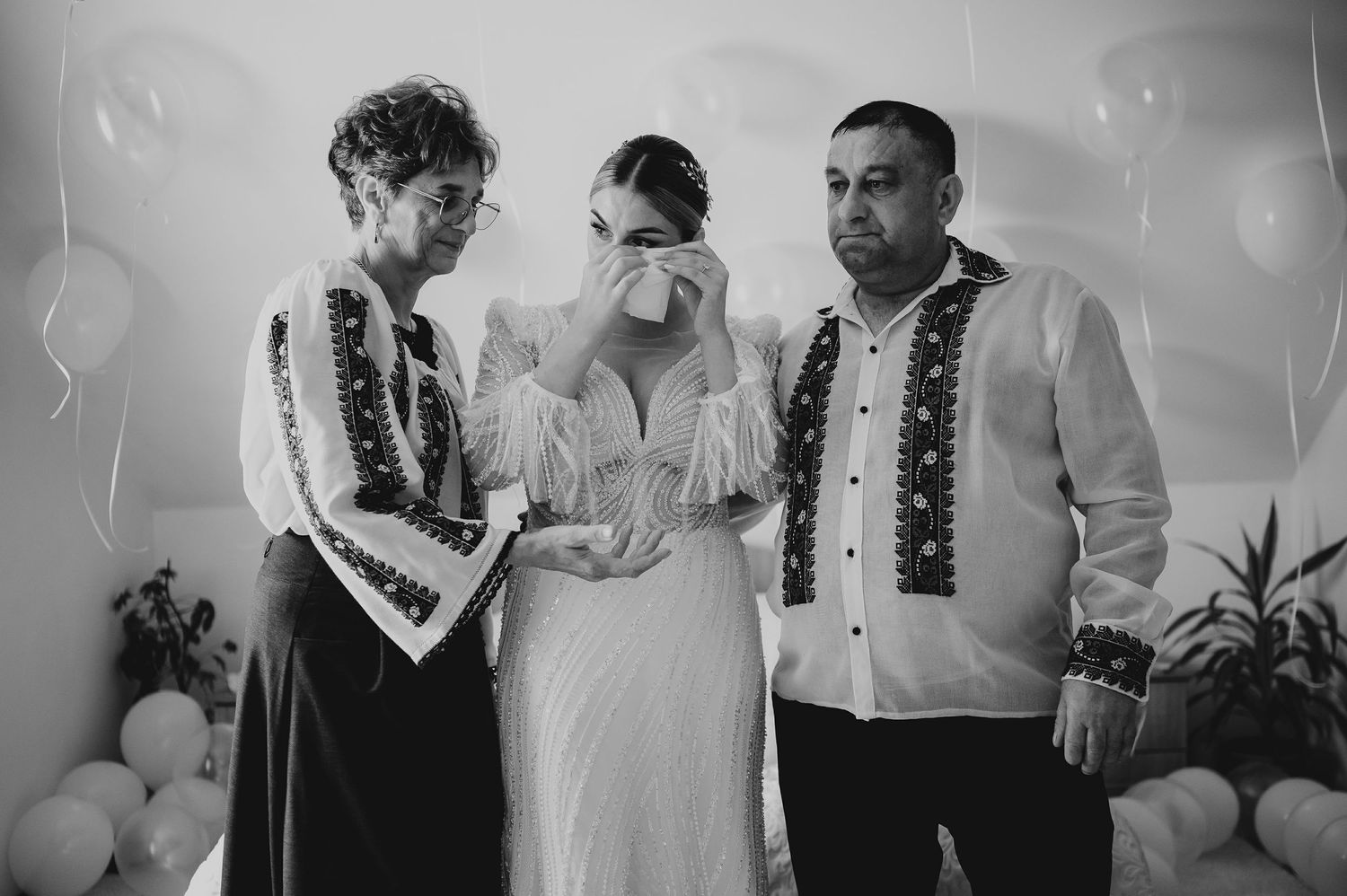 Black and white photo of emotional bride wiping tears flanked by parents in traditional embroidered Romanian attire.