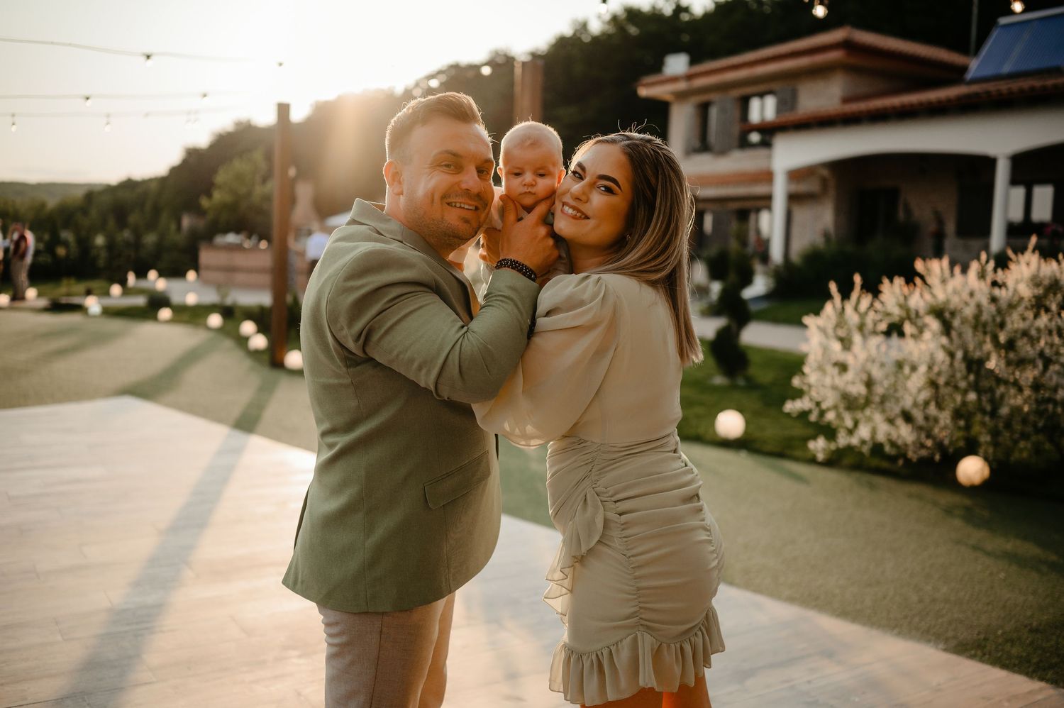 Happy family in green outfits holding baby while smiling at camera during golden hour outdoors.