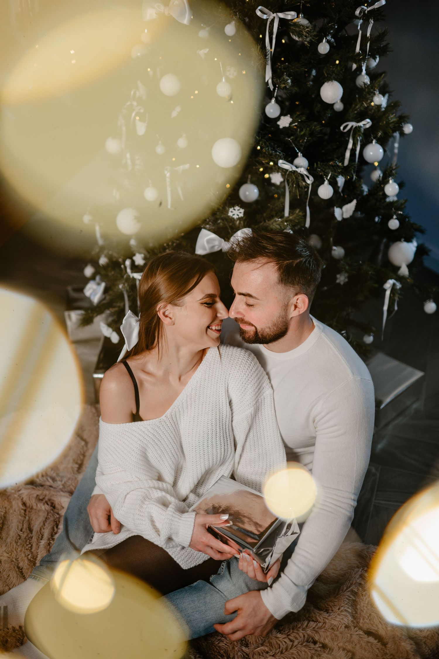Smiling couple embracing near a white-decorated Christmas tree with bokeh light effects.