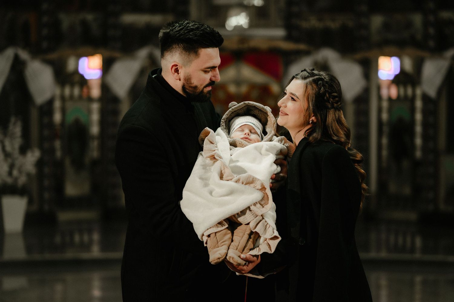 Parents in matching black outfits tenderly looking at newborn wrapped in white blanket.