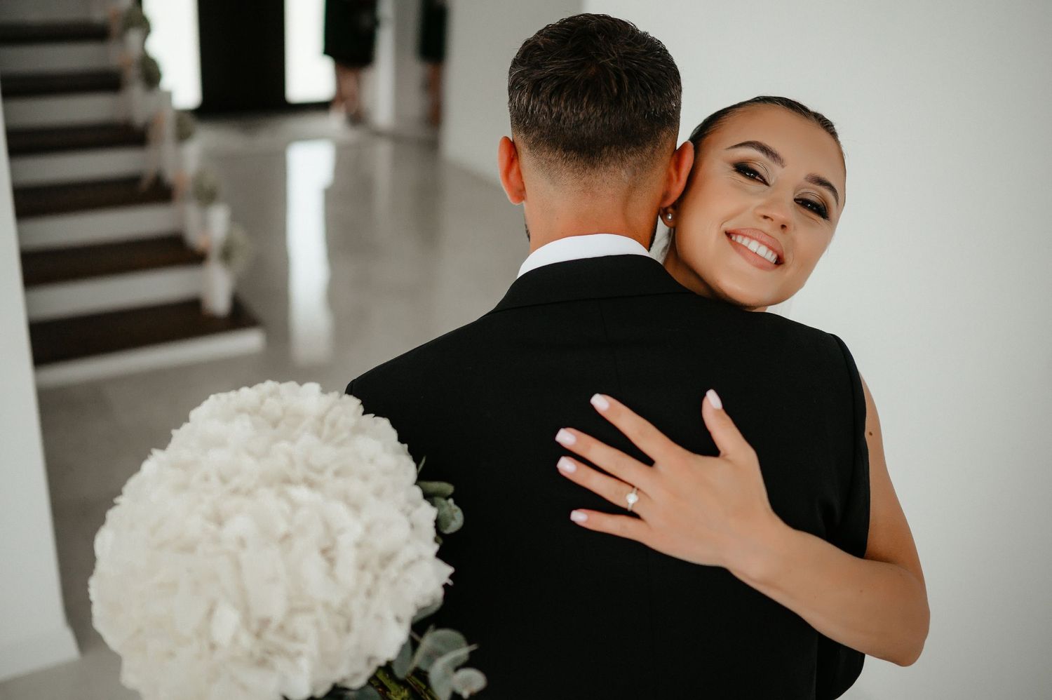 Smiling bride in white gown embraces groom from behind while holding a lush white floral bouquet.