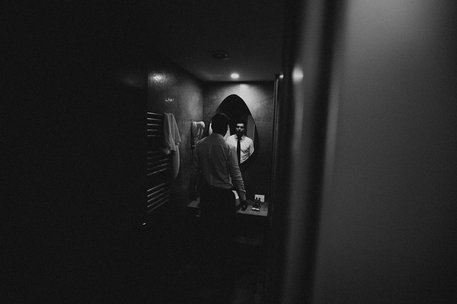 Groom preparing in dimly lit bathroom reflected in arched mirror before wedding ceremony.