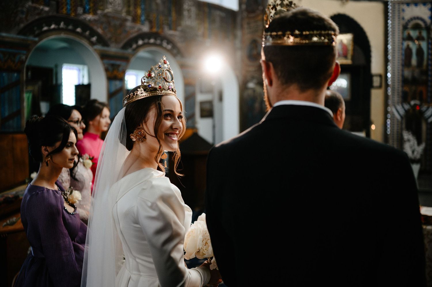 Smiling bride wearing ornate crown facing groom at Orthodox church wedding altar.