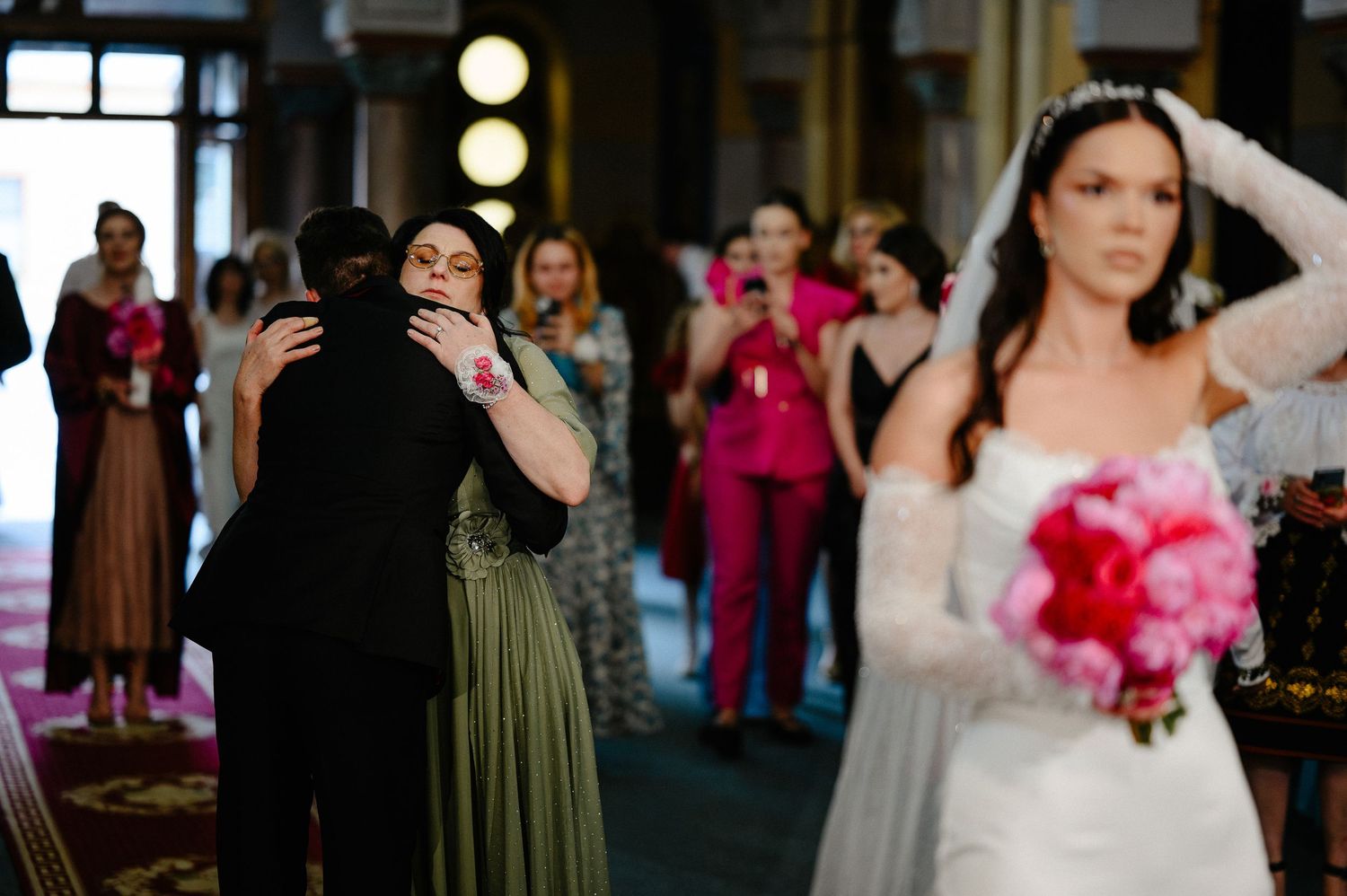 Emotional wedding guest hugs attendee while bride in white gown holds pink bouquet in foreground.