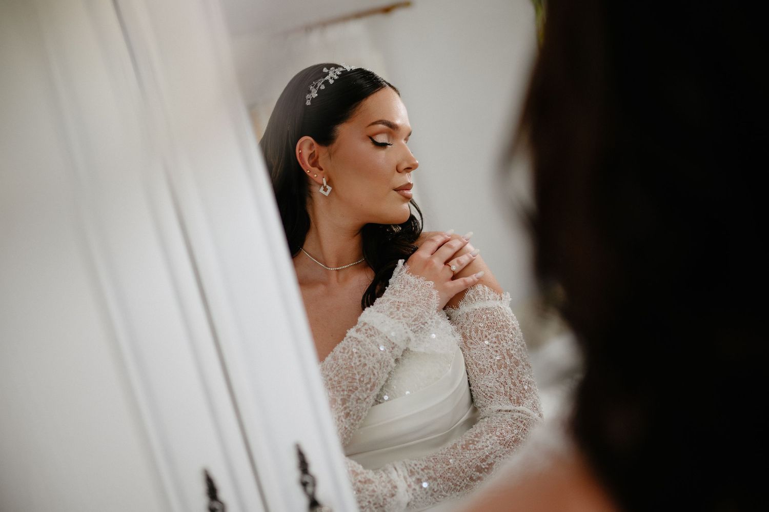Bride in lace long-sleeve wedding dress admiring her reflection in a mirror before ceremony.