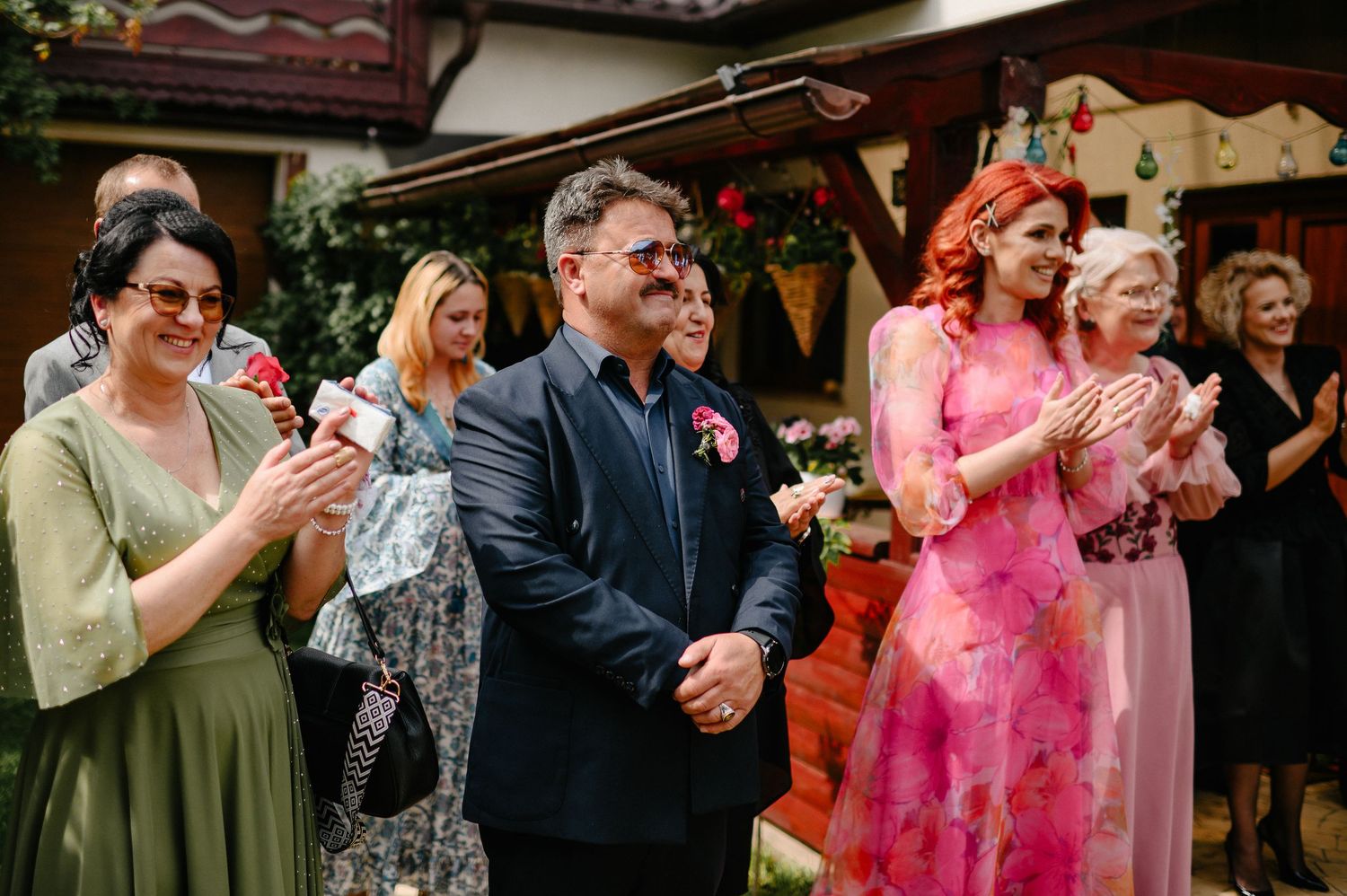 Smiling wedding guests applaud in colorful formal attire at an outdoor wedding celebration venue.