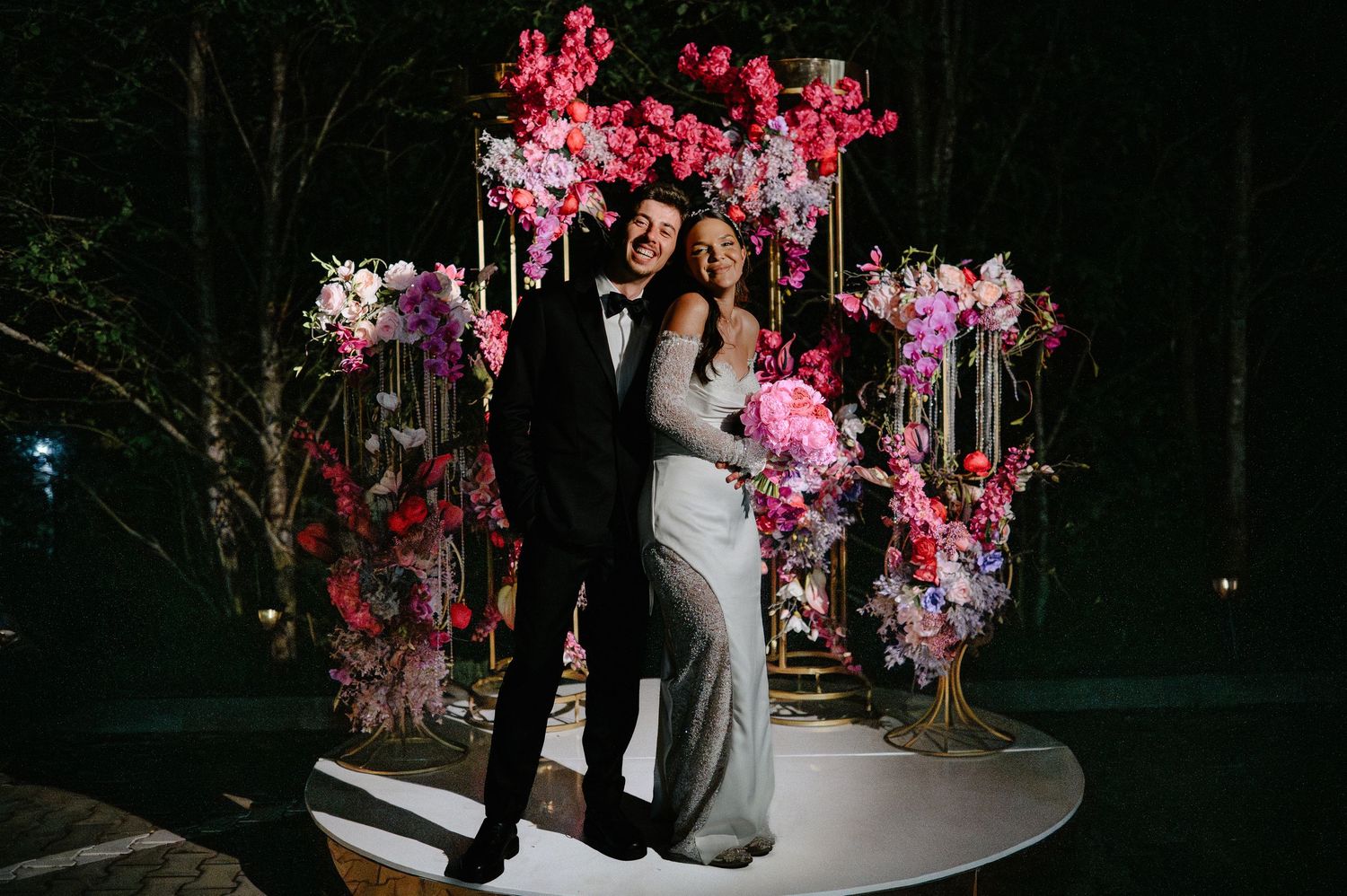 Happy newlyweds posing before stunning pink floral wedding arch with candles at nighttime ceremony.