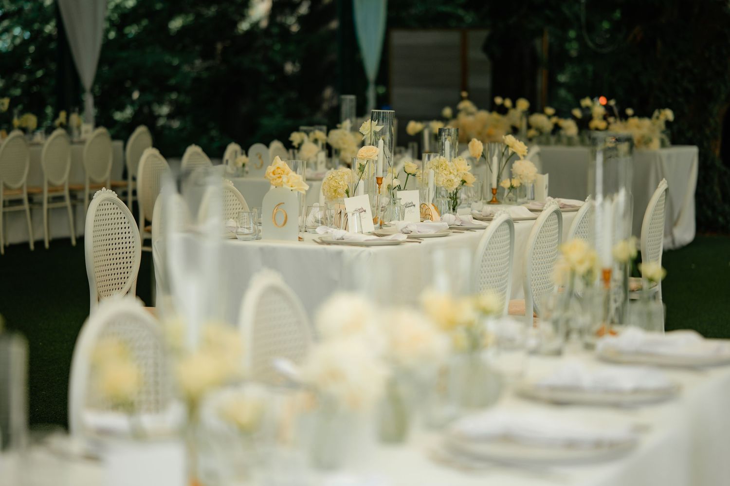Romantic outdoor wedding reception tables with white floral arrangements, wicker chairs, and soft yellow blooms.