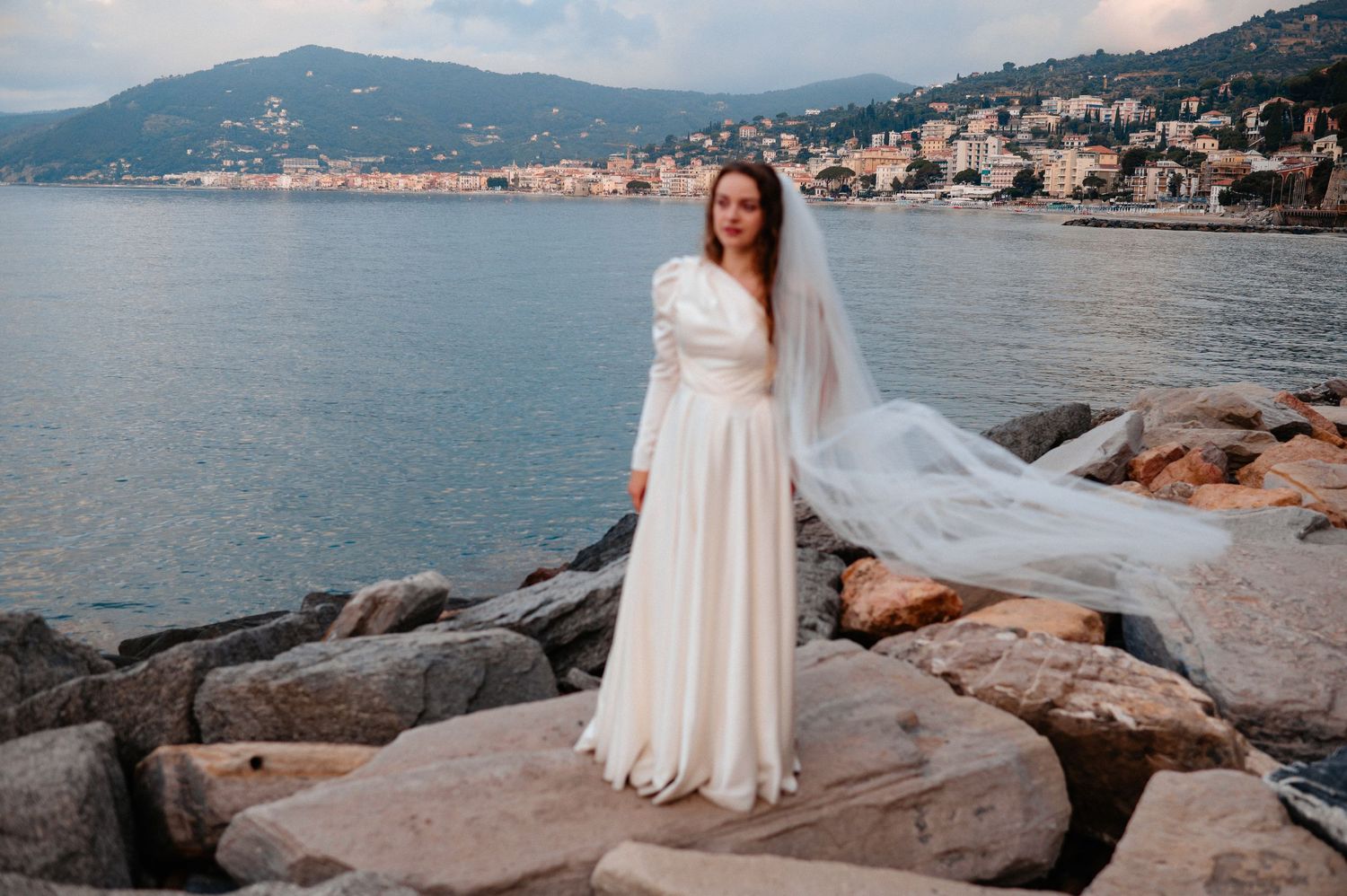 Bride in white gown and veil stands on rocky Mediterranean coastline with seaside town in background.