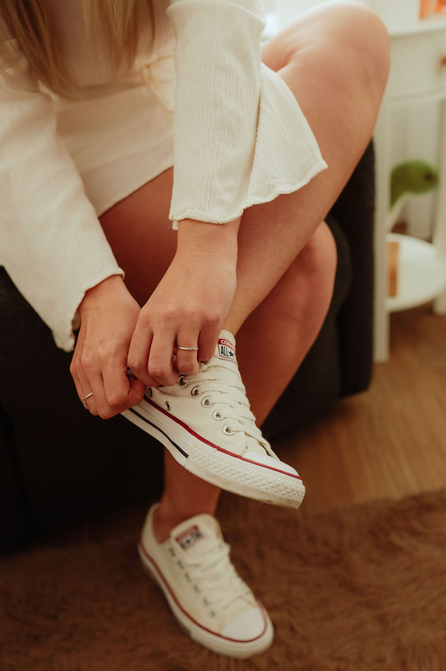 Woman putting on white Converse Chuck Taylor low-top sneakers while seated indoors.