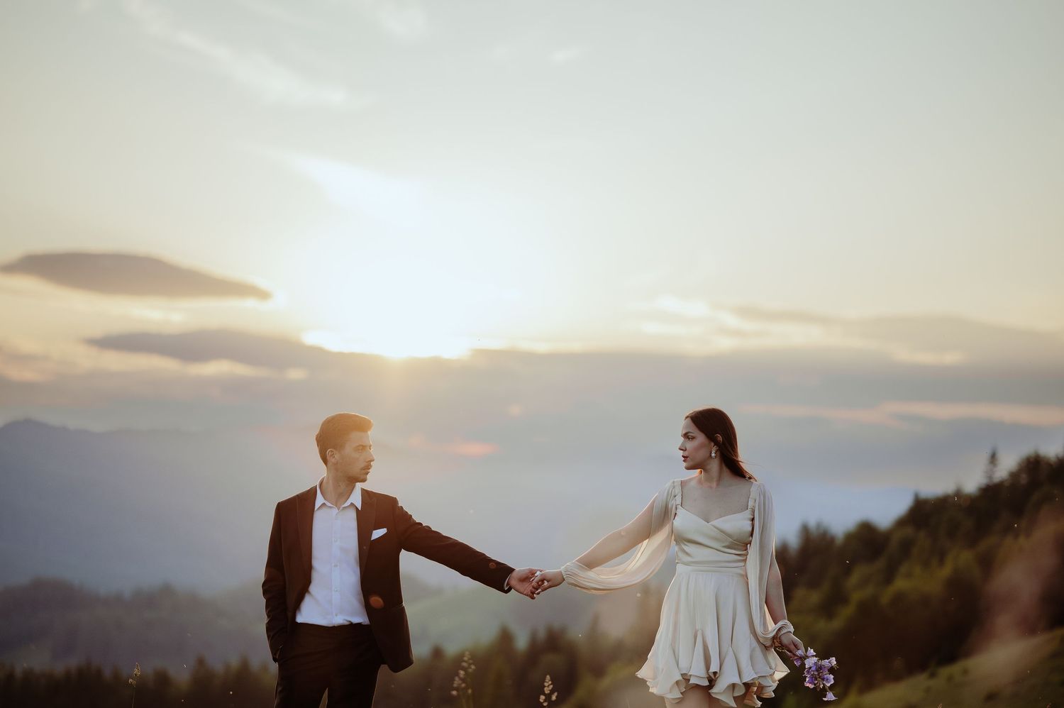 A bride and groom hold hands while walking through a mountain meadow during a stunning sunset.