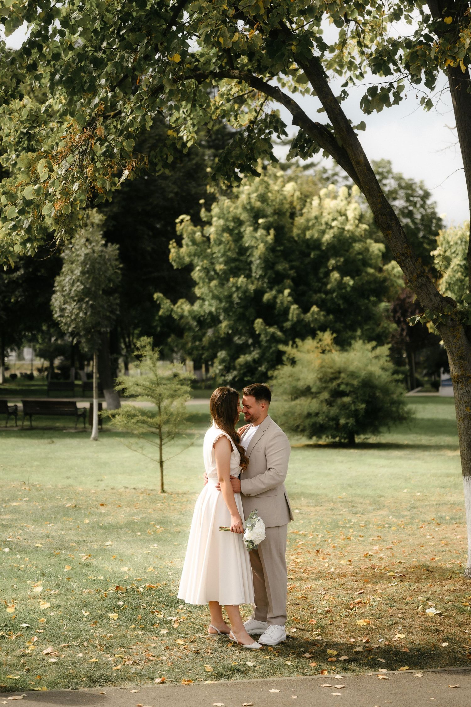 A couple in wedding attire shares a romantic moment in a lush green park, bride holding a bouquet.