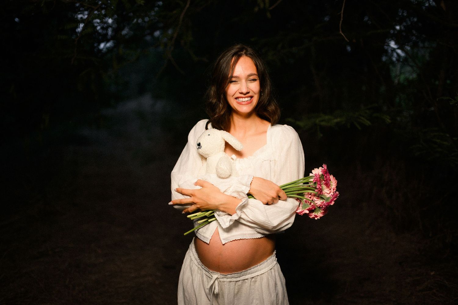 Smiling pregnant woman in white dress holding a white stuffed animal and pink flowers in a dark forest setting.