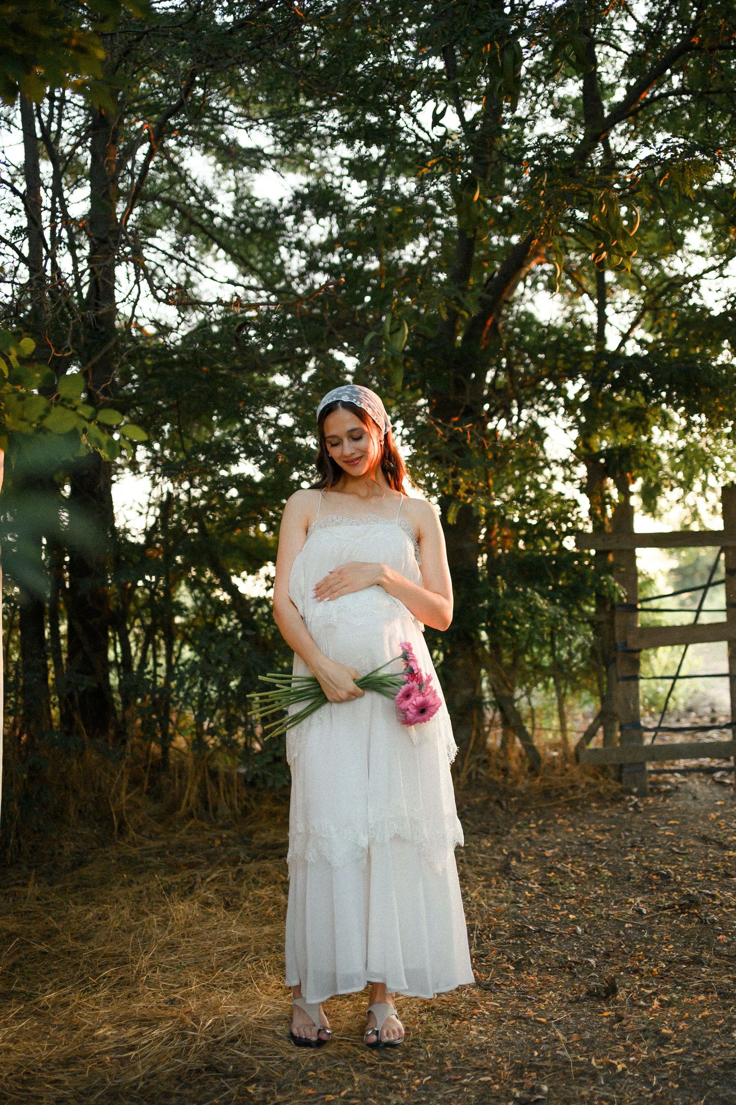 Pregnant woman in white dress holding pink flowers, standing outdoors near trees and wooden fence in golden sunlight.