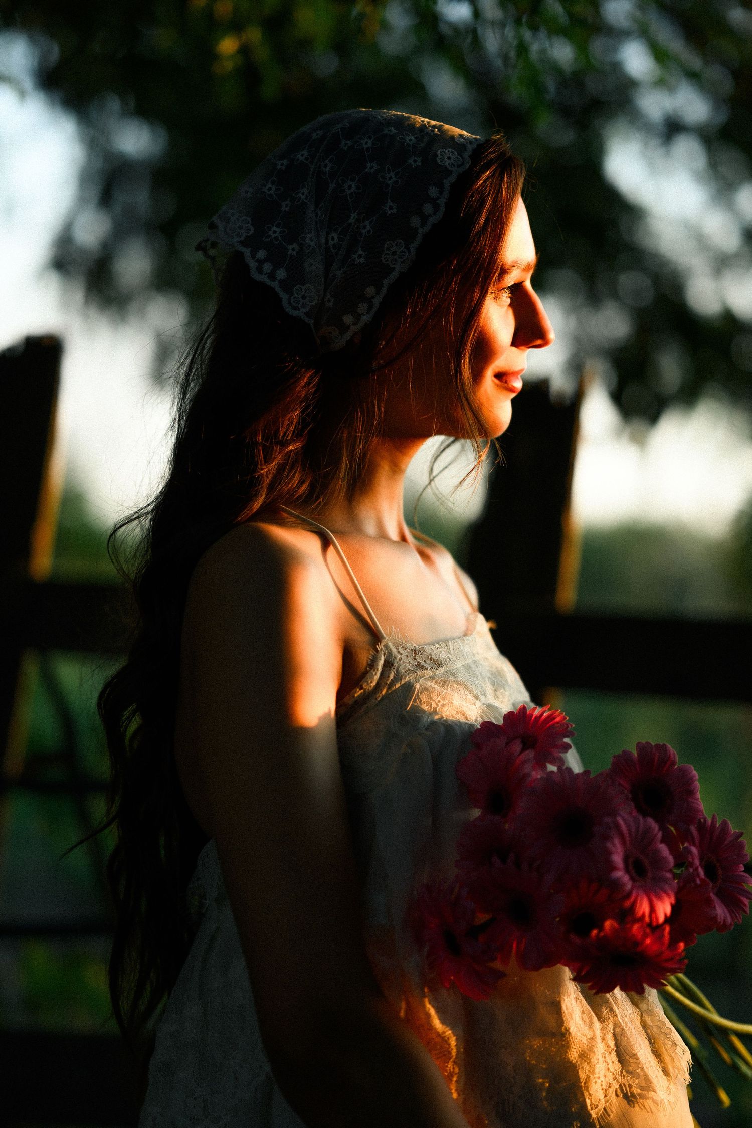 Young woman with long dark hair and headscarf holds red flowers in golden sunset light outdoors.