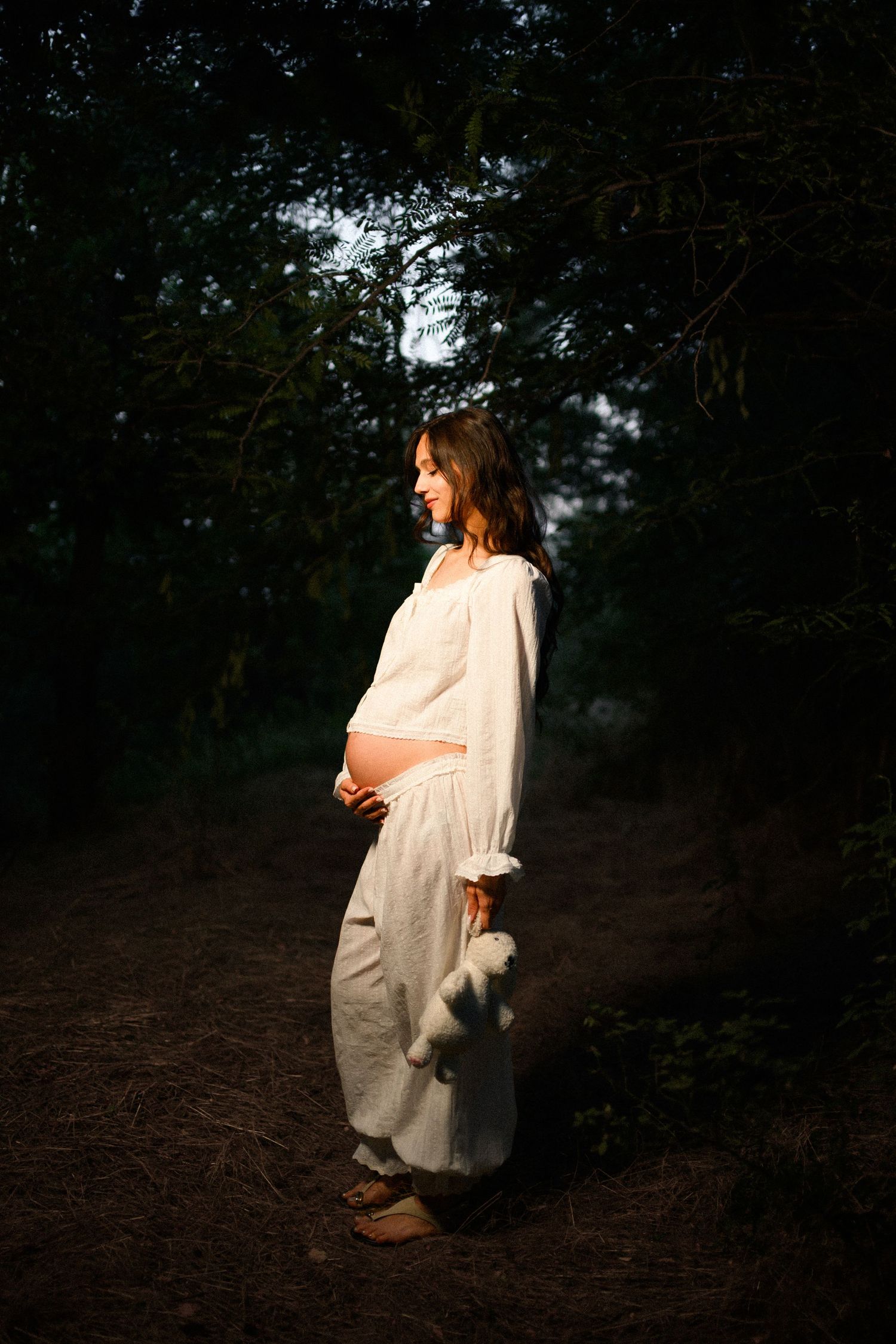 Pregnant woman in white dress standing in sunlit forest, cradling baby bump, serene maternity photo.