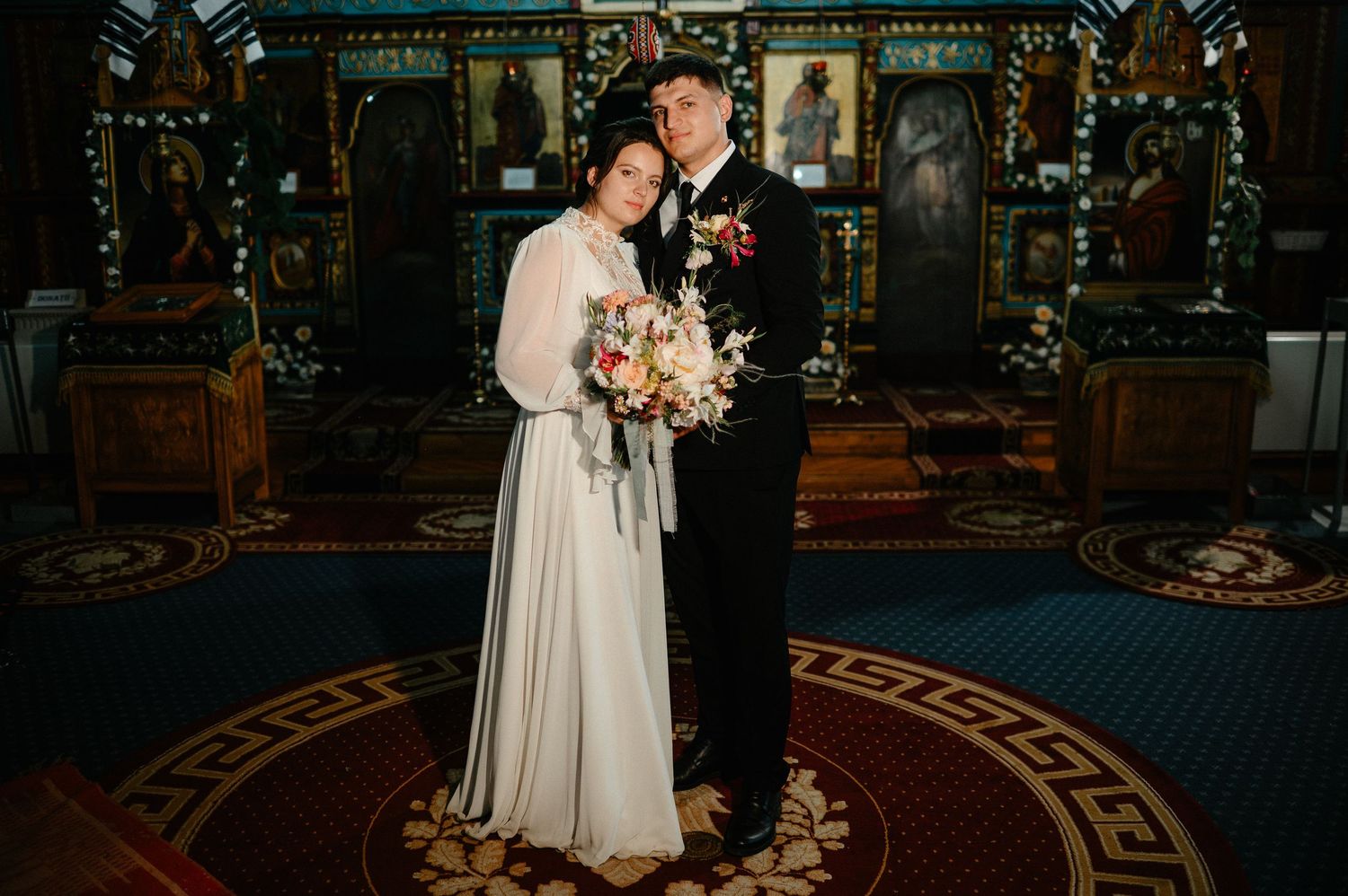 Bride and groom pose together inside an ornate Orthodox church with decorative icons and colorful interior.