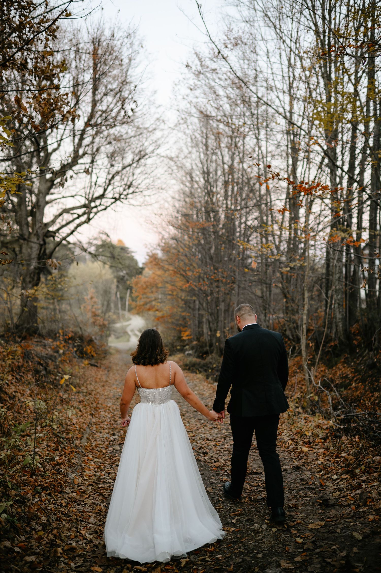 Bride and groom walking hand in hand down a leaf-covered forest path in autumn, bride wearing a white gown.