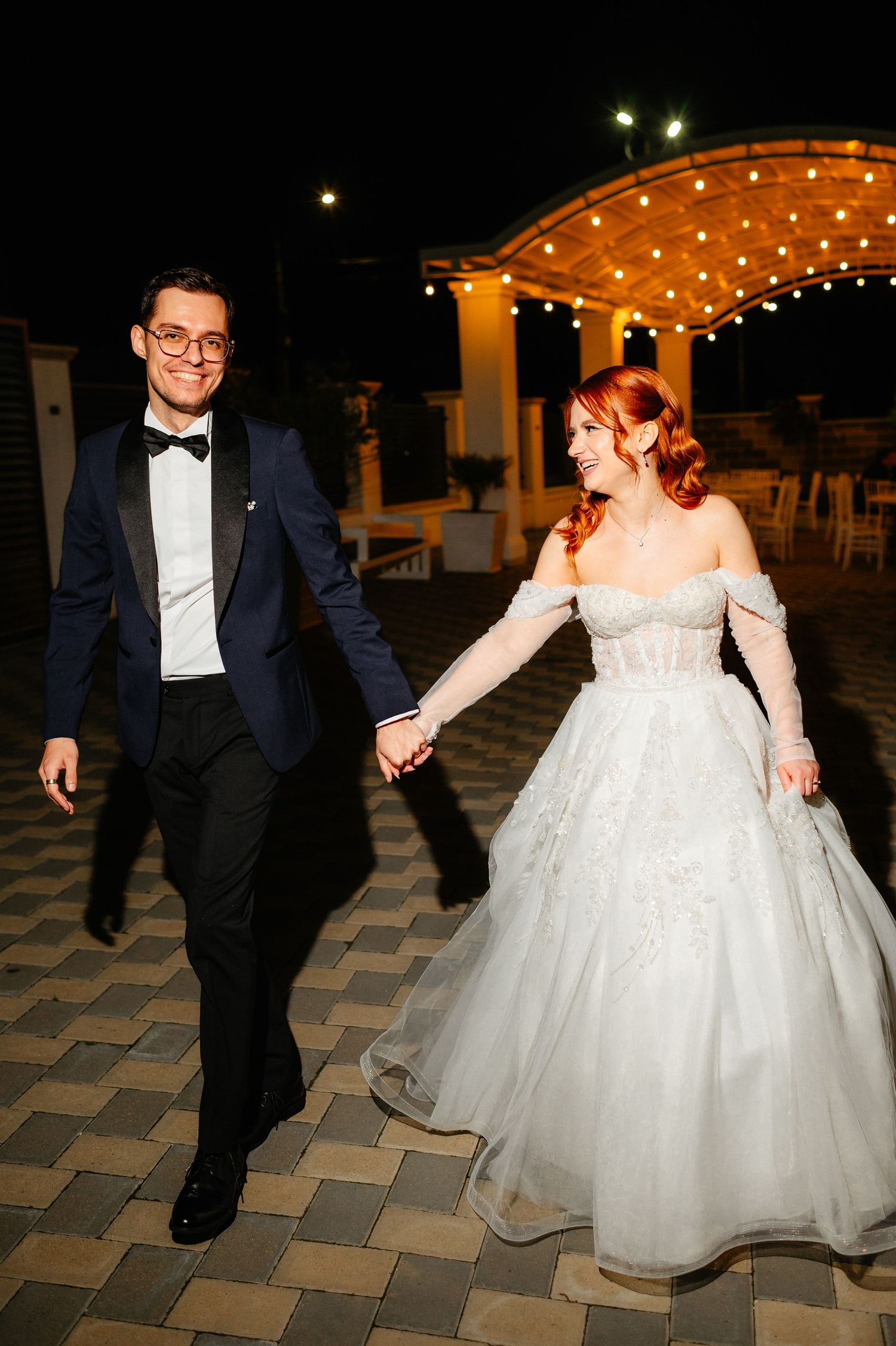 Bride in off-shoulder lace gown and groom in navy tuxedo hold hands walking at outdoor night wedding venue.