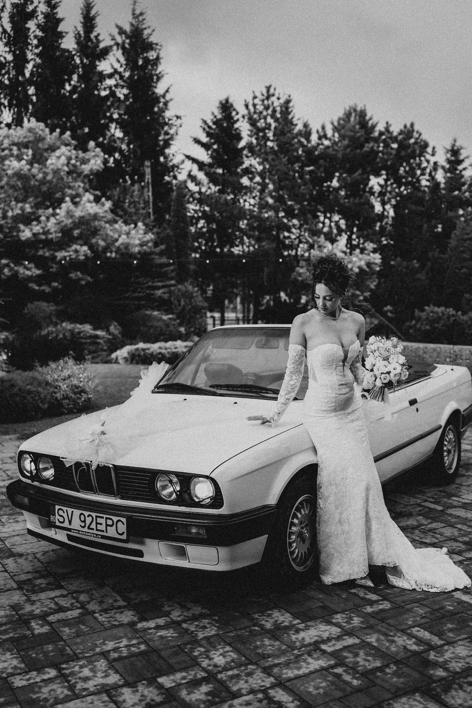 Bride in lace wedding gown poses beside a classic BMW convertible adorned with ribbon in a black and white photo.