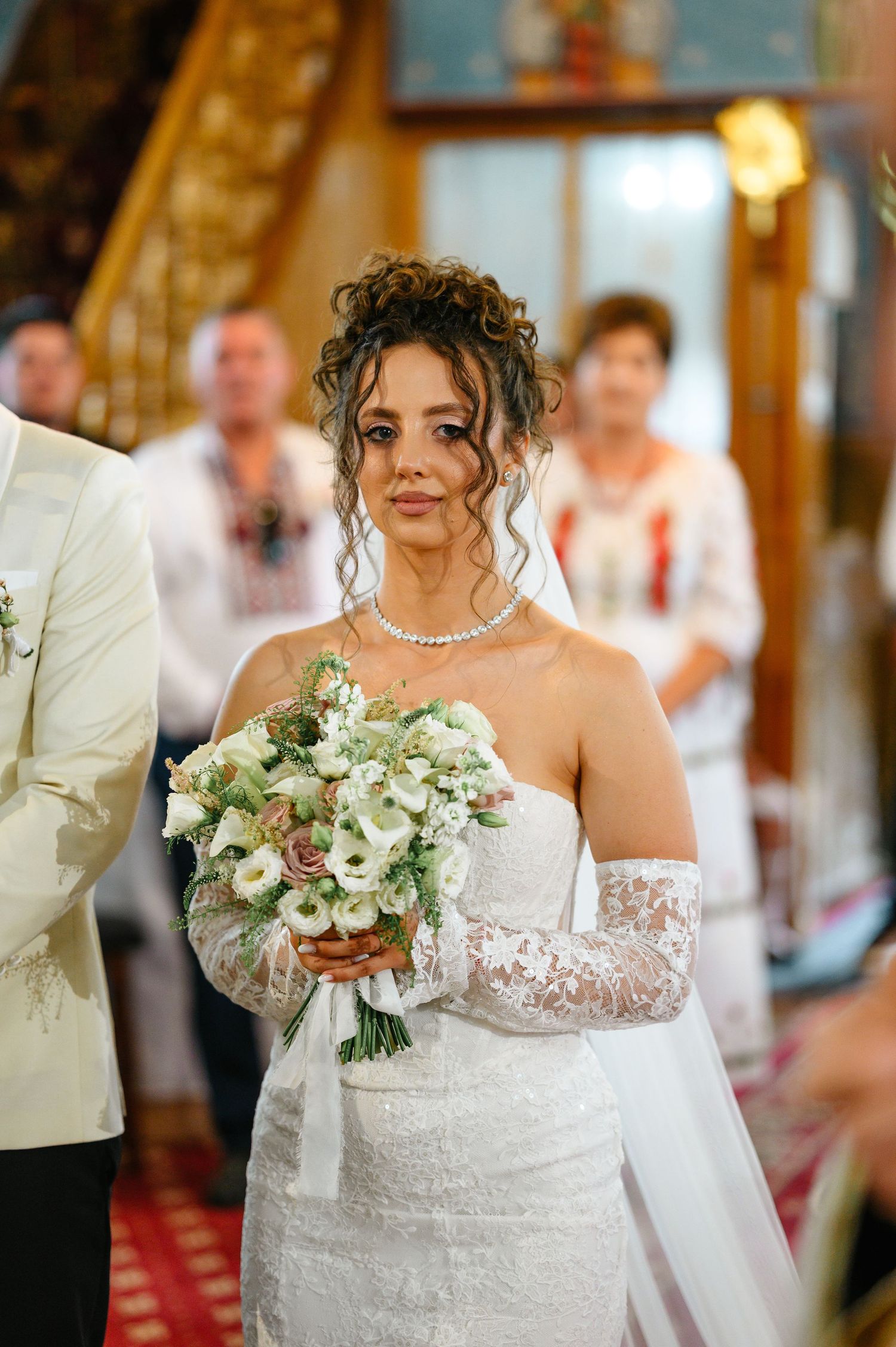 Elegant bride in white lace gown holding bouquet of white flowers at church wedding ceremony.