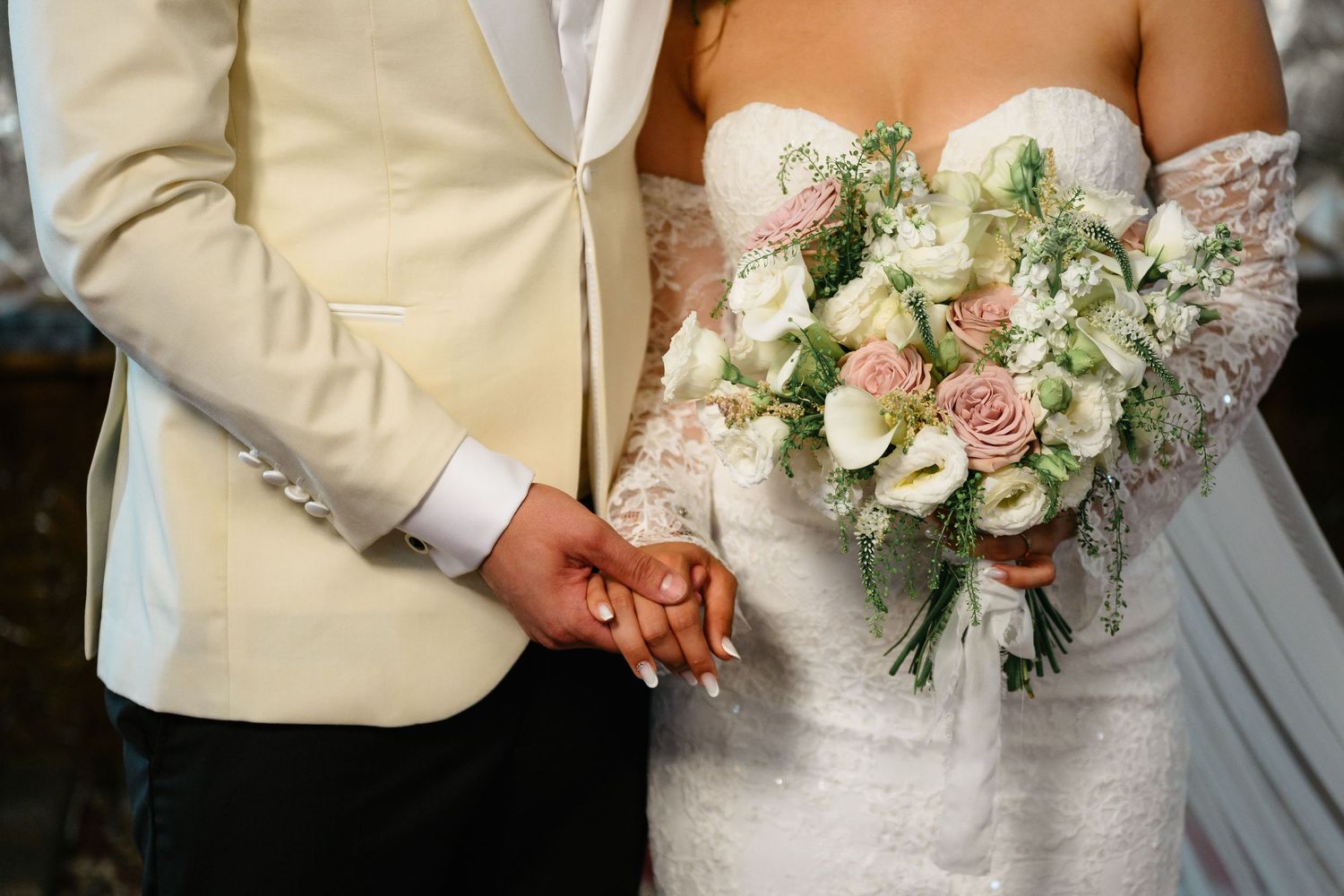 Interracial couple holding hands at wedding, bride in lace gown holding pink and white bouquet, groom in cream suit.