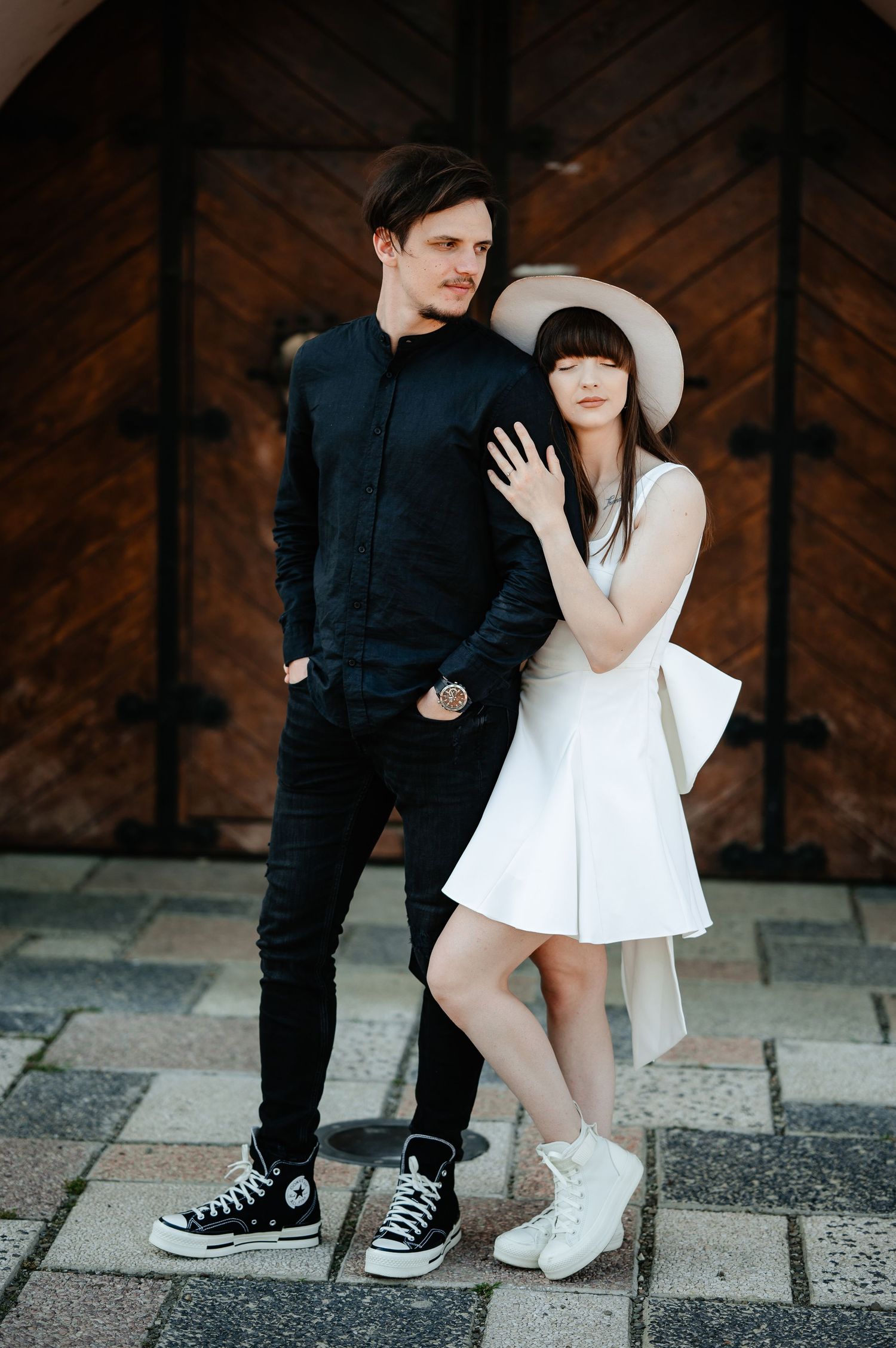 Stylish couple posing together, man in black outfit and woman in white dress with wide-brim hat.