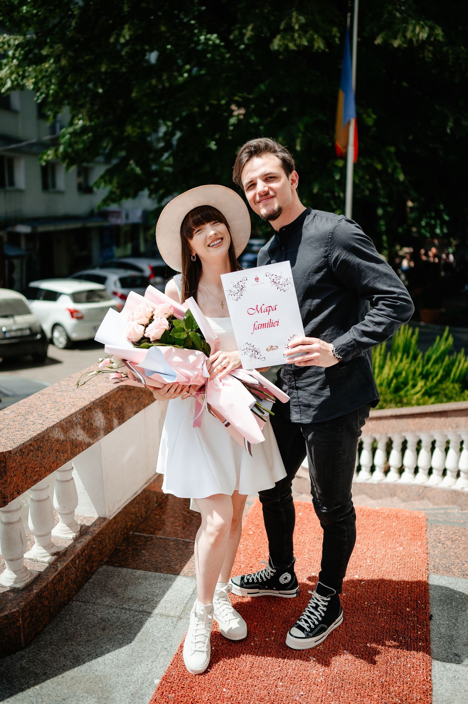Newlyweds pose outdoors holding marriage certificate and pink bouquet, bride in white dress and hat, groom in black.