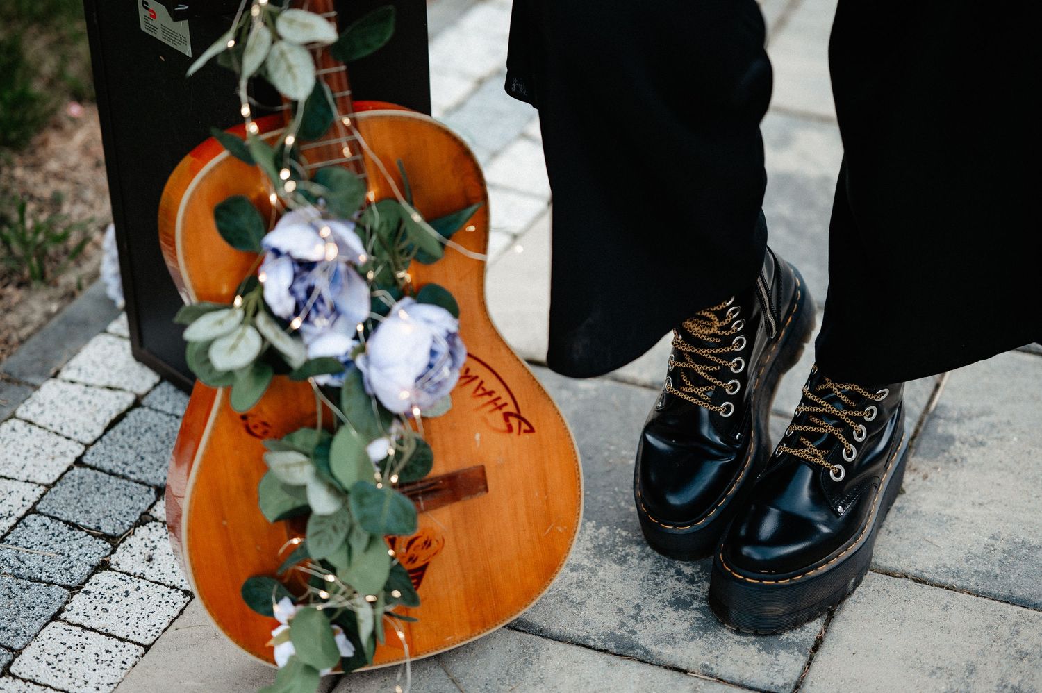 Acoustic guitar with blue floral arrangement leaning against a wall beside black lace-up platform boots.