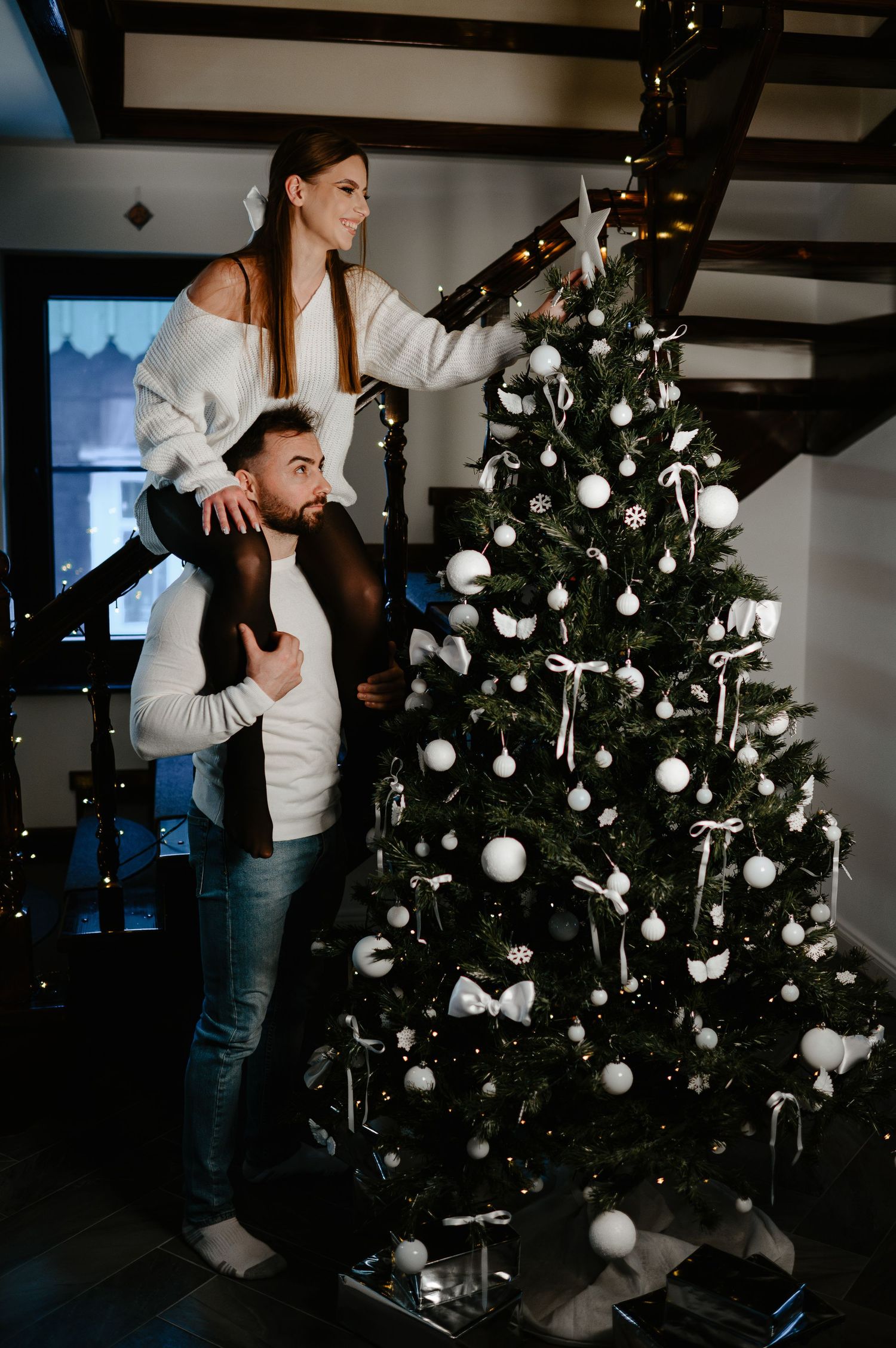 Woman sitting on man's shoulders to place a star on top of a Christmas tree.