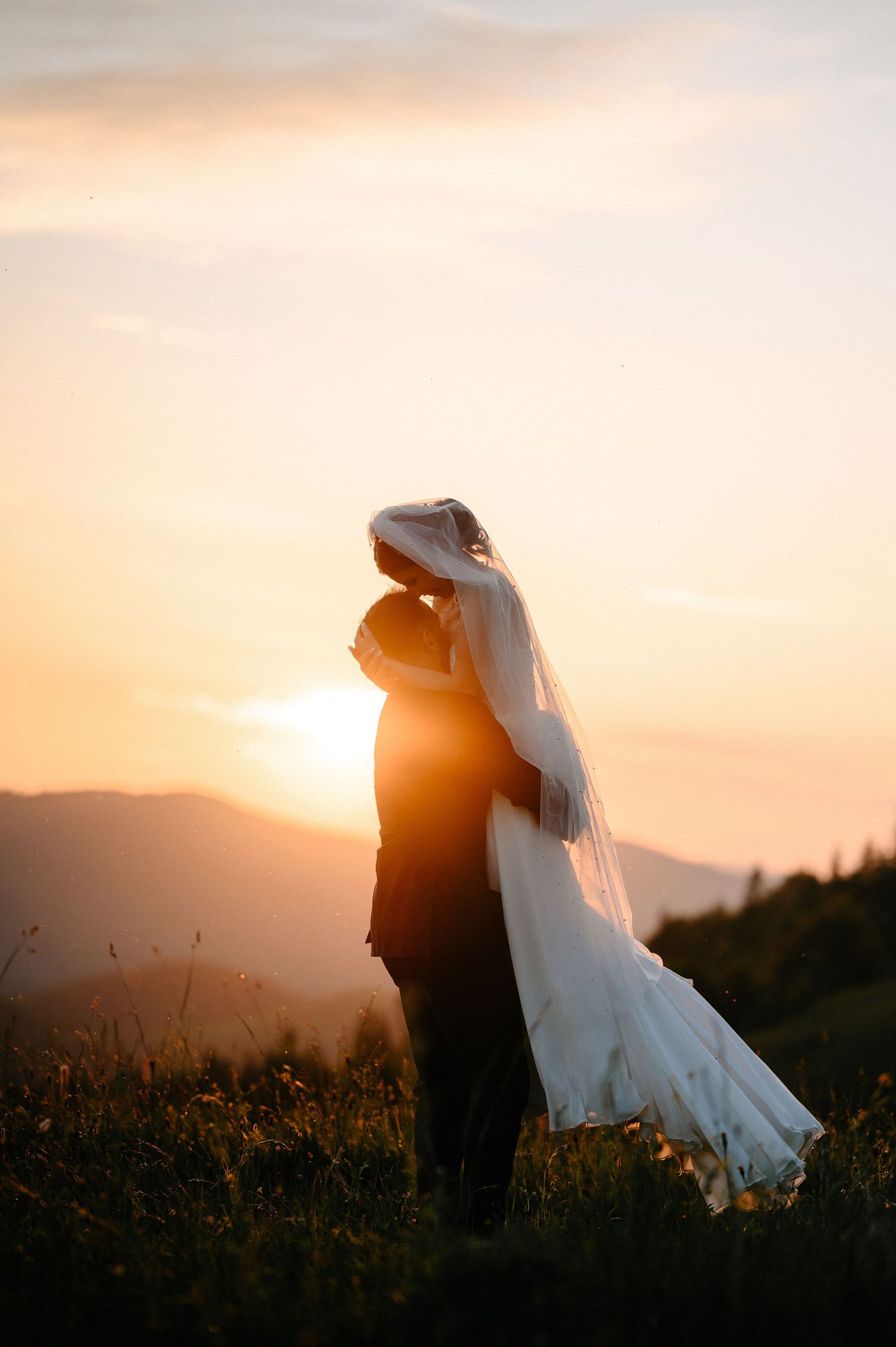 Bride and groom silhouetted against a breathtaking golden sunset in a romantic mountain meadow embrace.