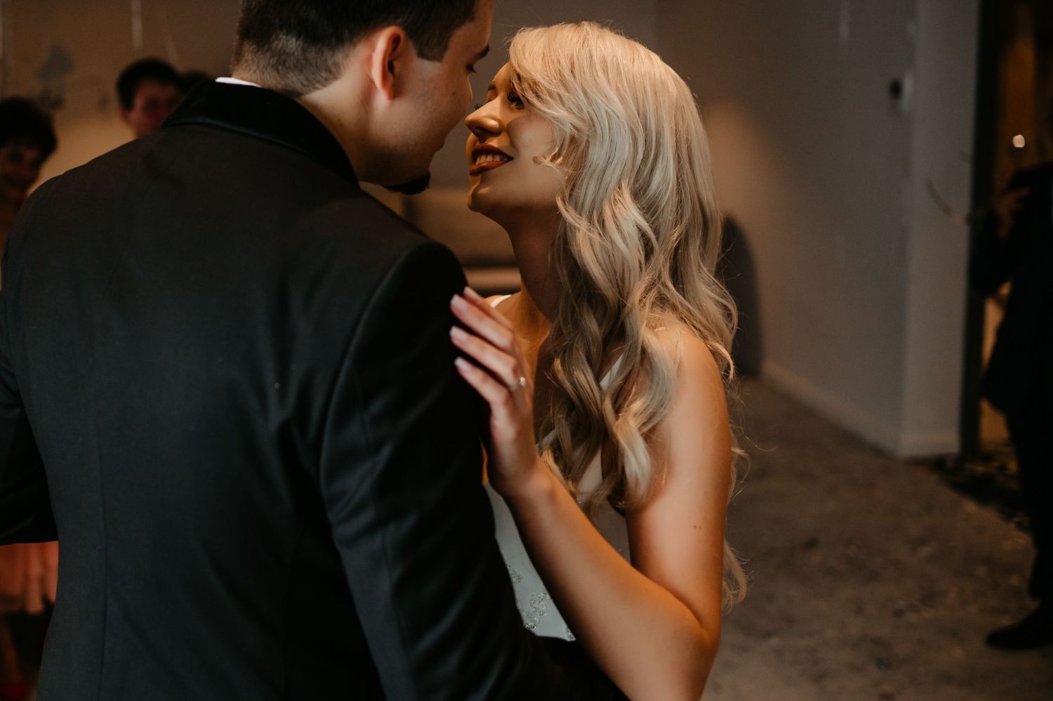 Bride with long blonde wavy hair gazing up at groom in black suit at wedding reception.