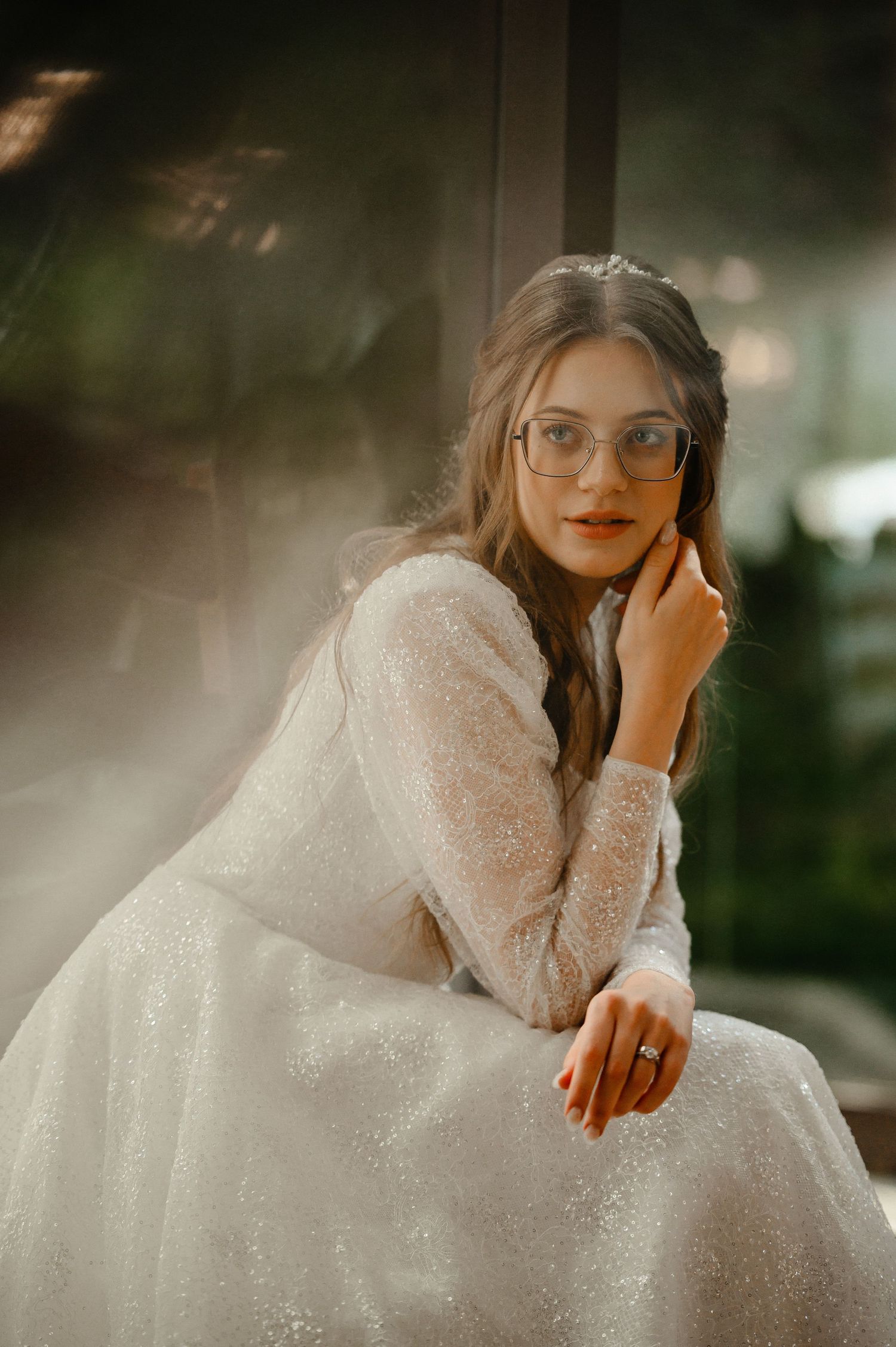 Elegant bride in sparkling white gown and glasses posing thoughtfully near a window.
