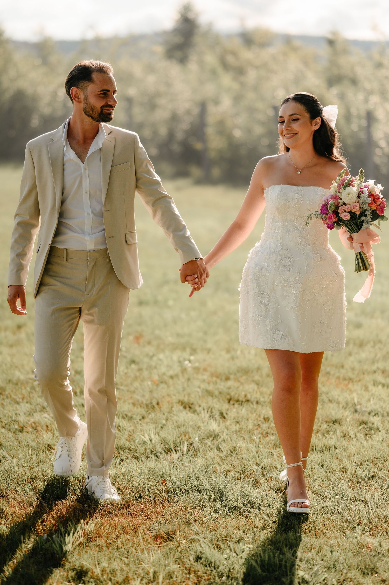 Bride in short lace dress and groom in beige suit hold hands walking through green meadow.