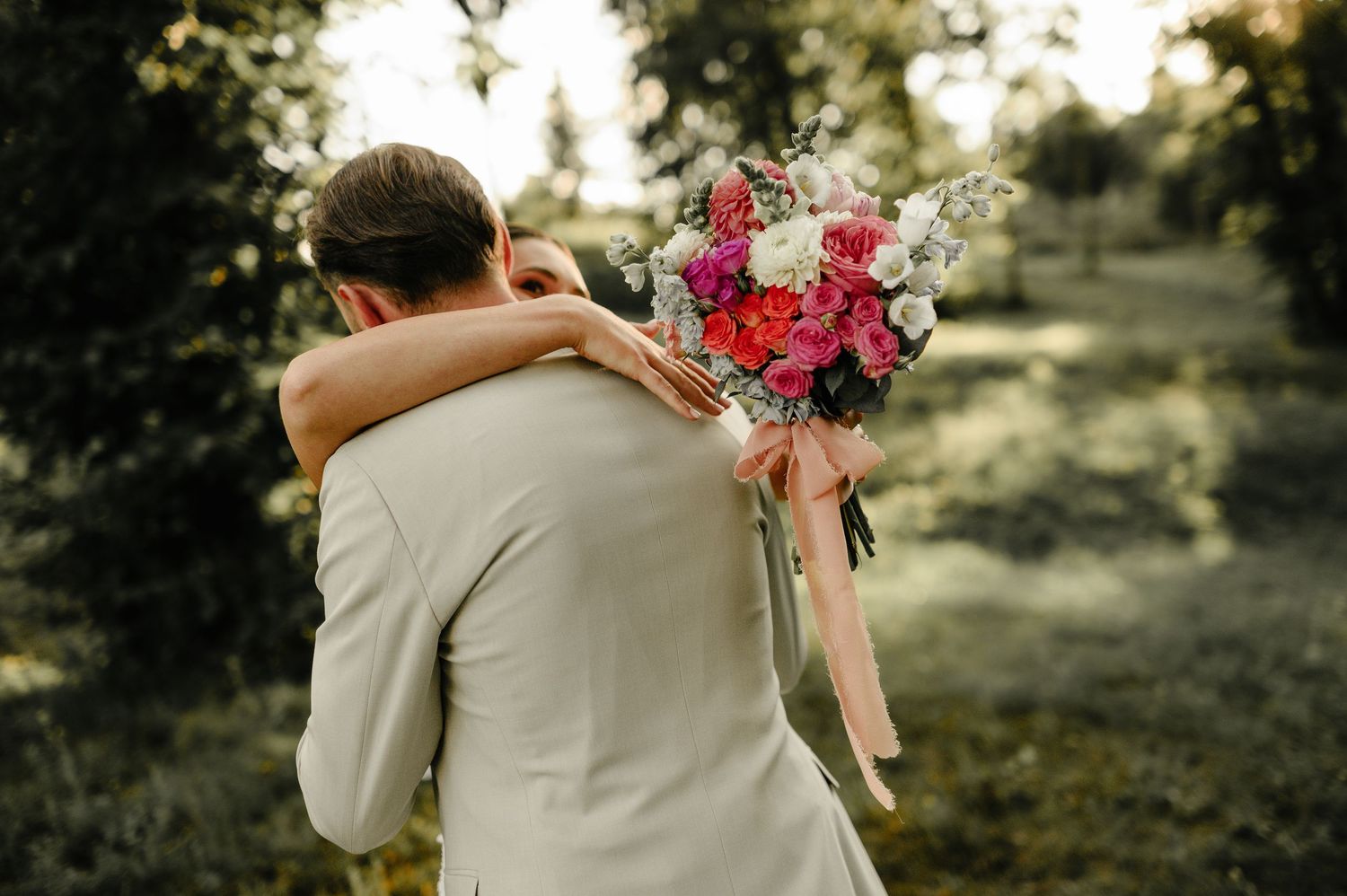 Bride and groom share a romantic embrace outdoors, bride holding a vibrant pink and white wedding bouquet.