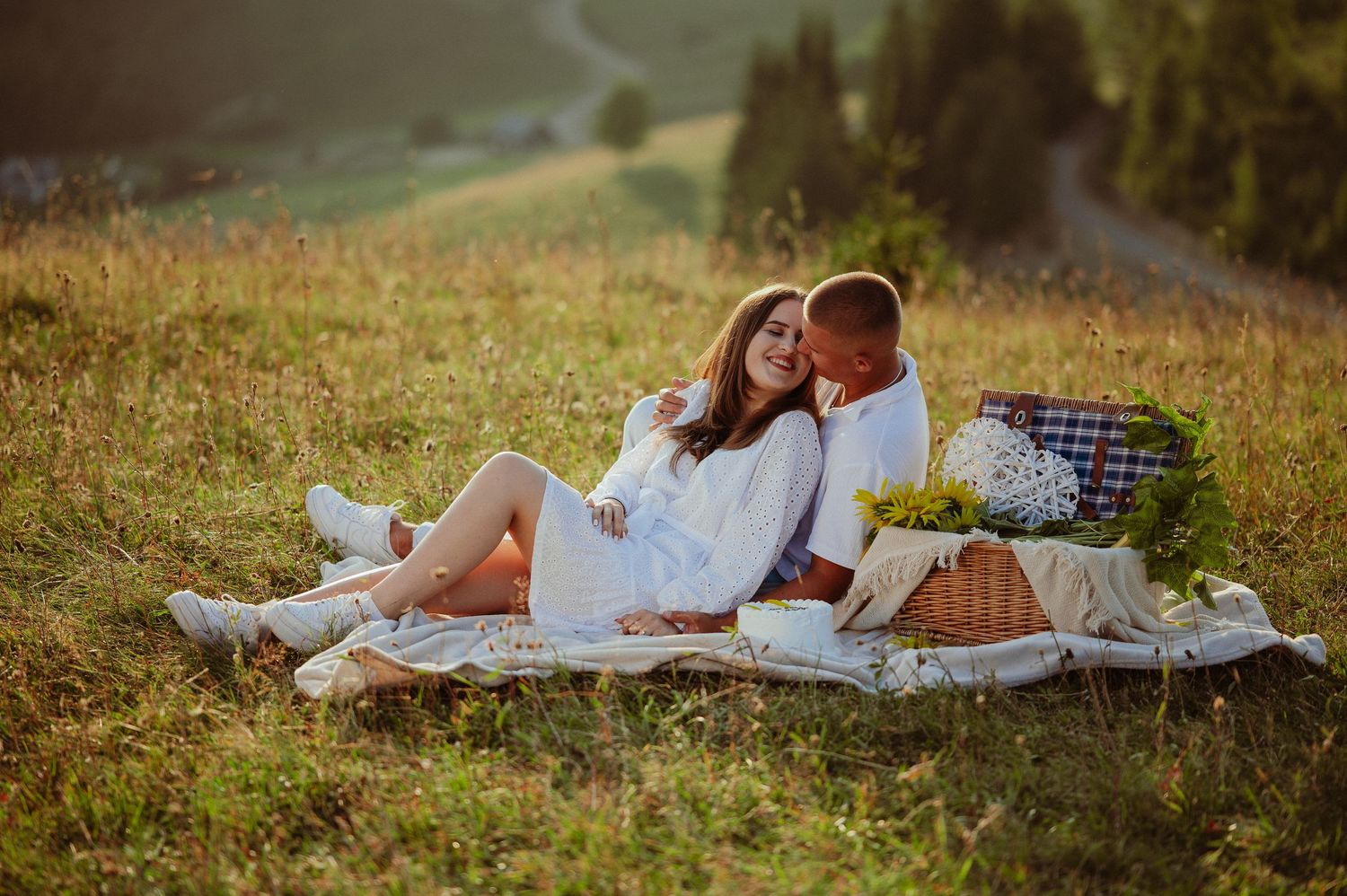 Couple in white outfits relaxing on picnic blanket in golden sunlit mountain meadow.