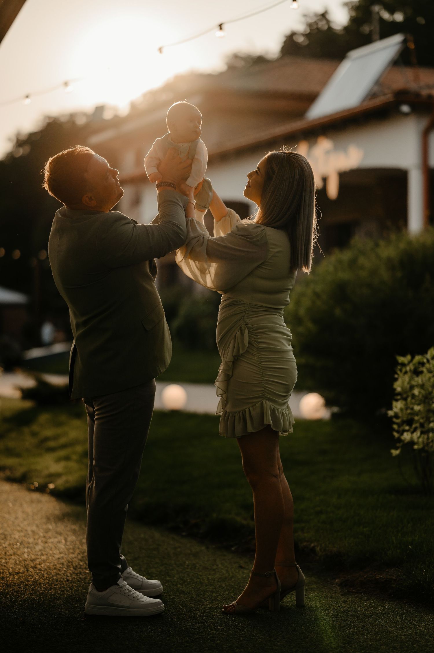 Family in coordinating green outfits lifting their baby together in golden sunset garden light.