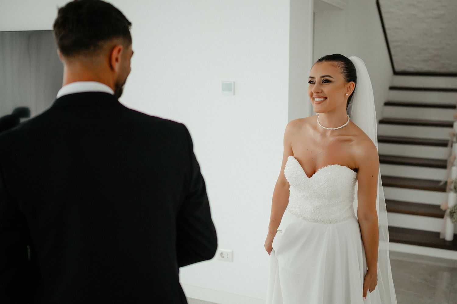 Bride in strapless wedding dress smiles warmly at groom during emotional first look indoors.