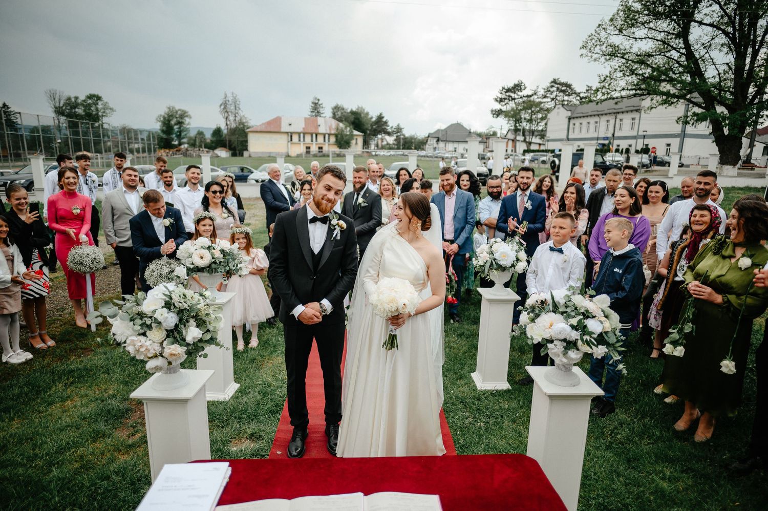 Bride and groom stand at outdoor wedding altar on red carpet surrounded by guests.
